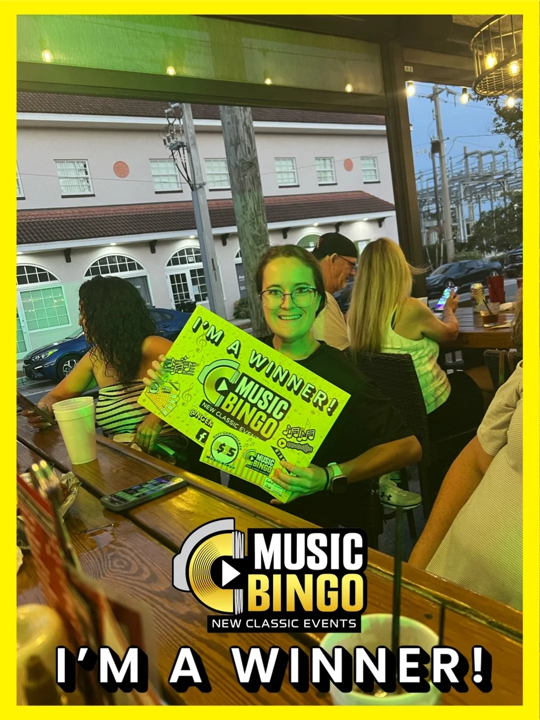 A woman sitting in a restaurant holding a sign that reads 'I'm a winner! Music Bingo'. She is smiling and wearing glasses. Other people are sitting at the table behind her, and the restaurant has large windows with a view of parked cars and a buildin