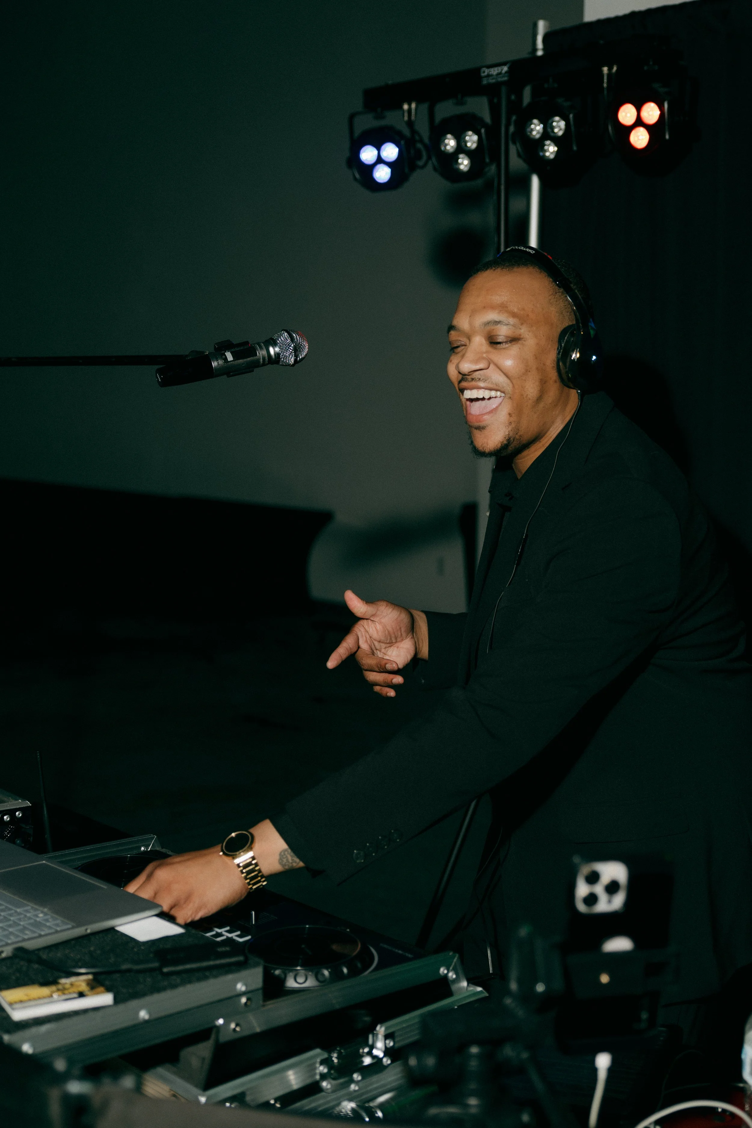 A man laughing and singing into a microphone while playing keyboard in a dark room with DJ lighting equipment in the background.