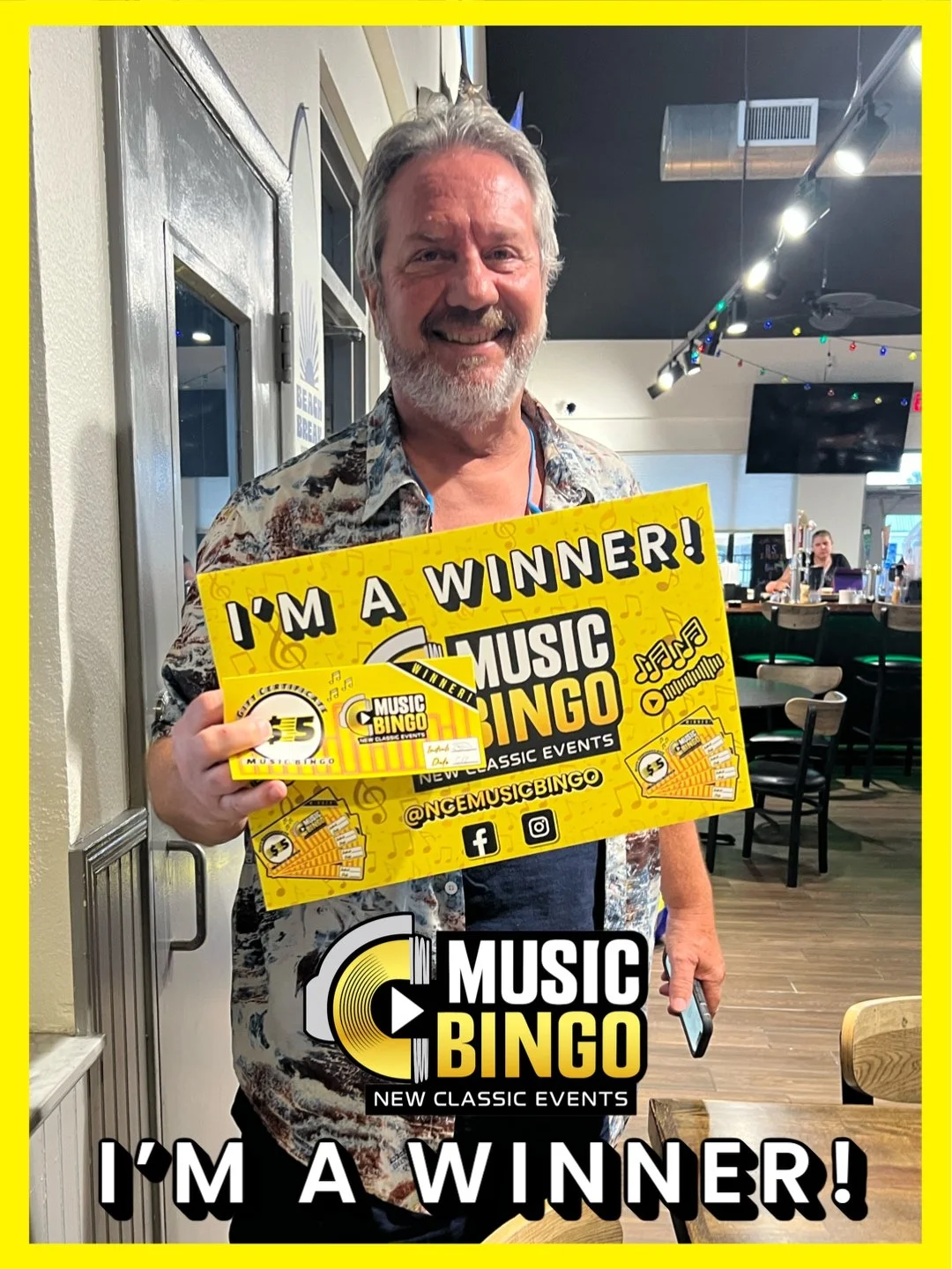 A smiling man with gray hair and beard holding a yellow 'Music Bingo' winner's card and a ticket, standing in a restaurant or bar. Behind him are tables, chairs, and a TV screen, with colorful string lights overhead.