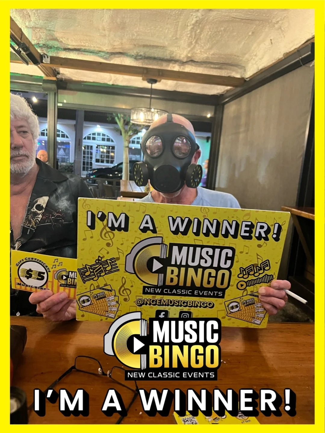 Person wearing a gas mask holding a yellow sign for Music Bingo at a table in a restaurant or bar with large windows and illuminated exterior.