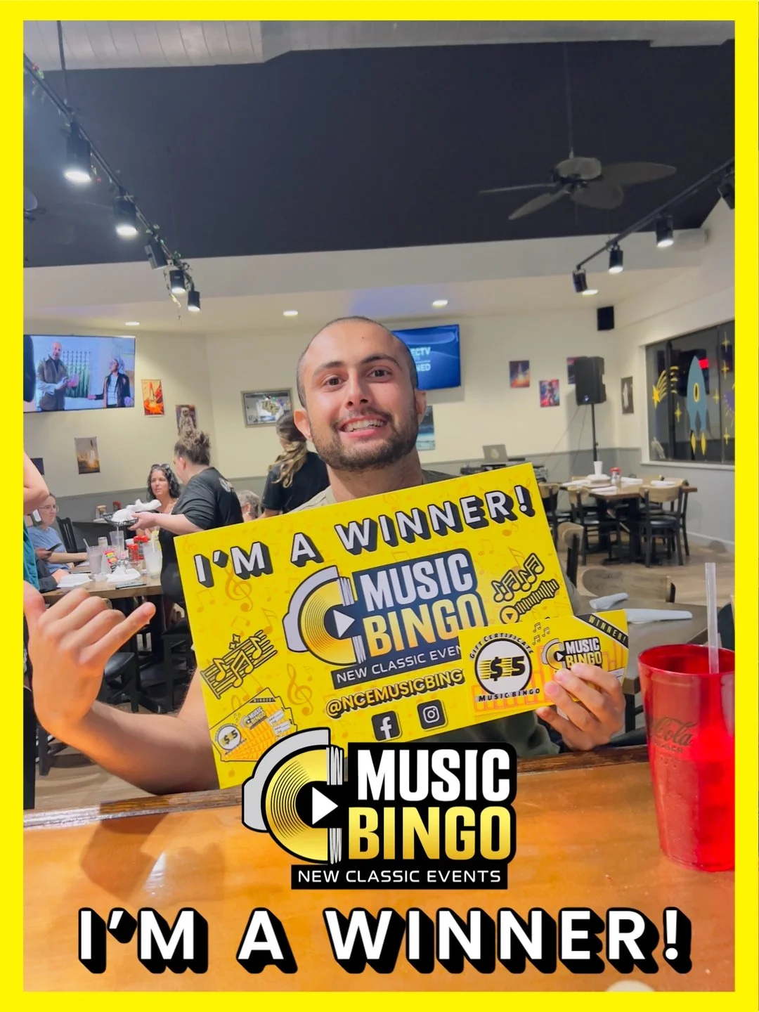 A smiling man holding a yellow 'Music Bingo' sign that says 'I'm a Winner!' inside a restaurant, with other patrons visible in the background.