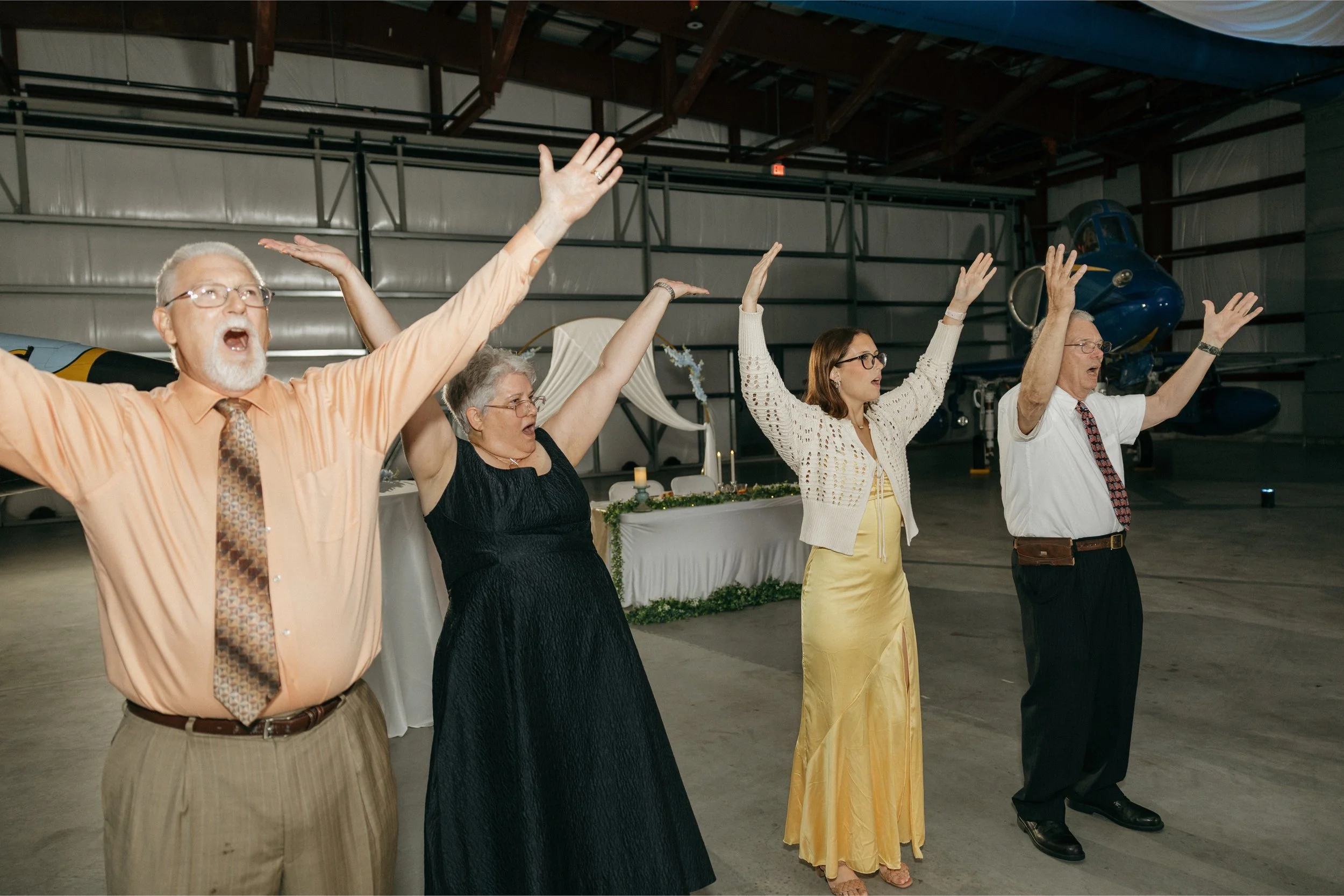 Four people, two men and two women, stand with arms raised in a large hangar, with a vintage blue airplane in the background, possibly during a celebration or event.