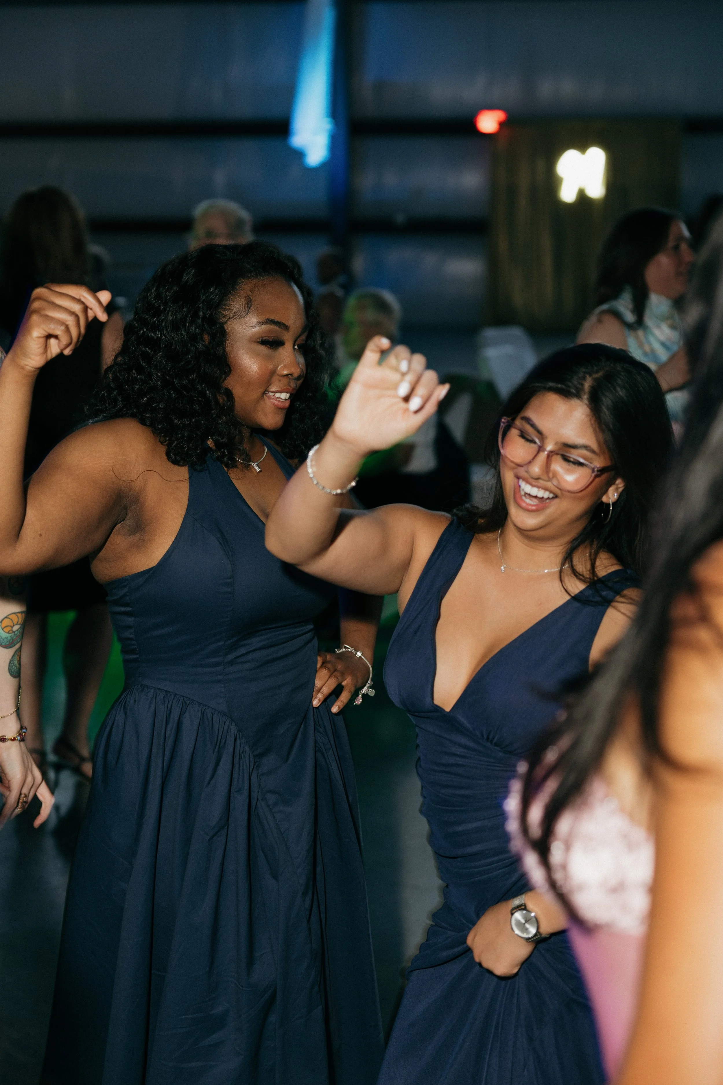 Two women in navy blue dresses dancing and smiling at a social event, surrounded by other attendees in a dimly lit room.