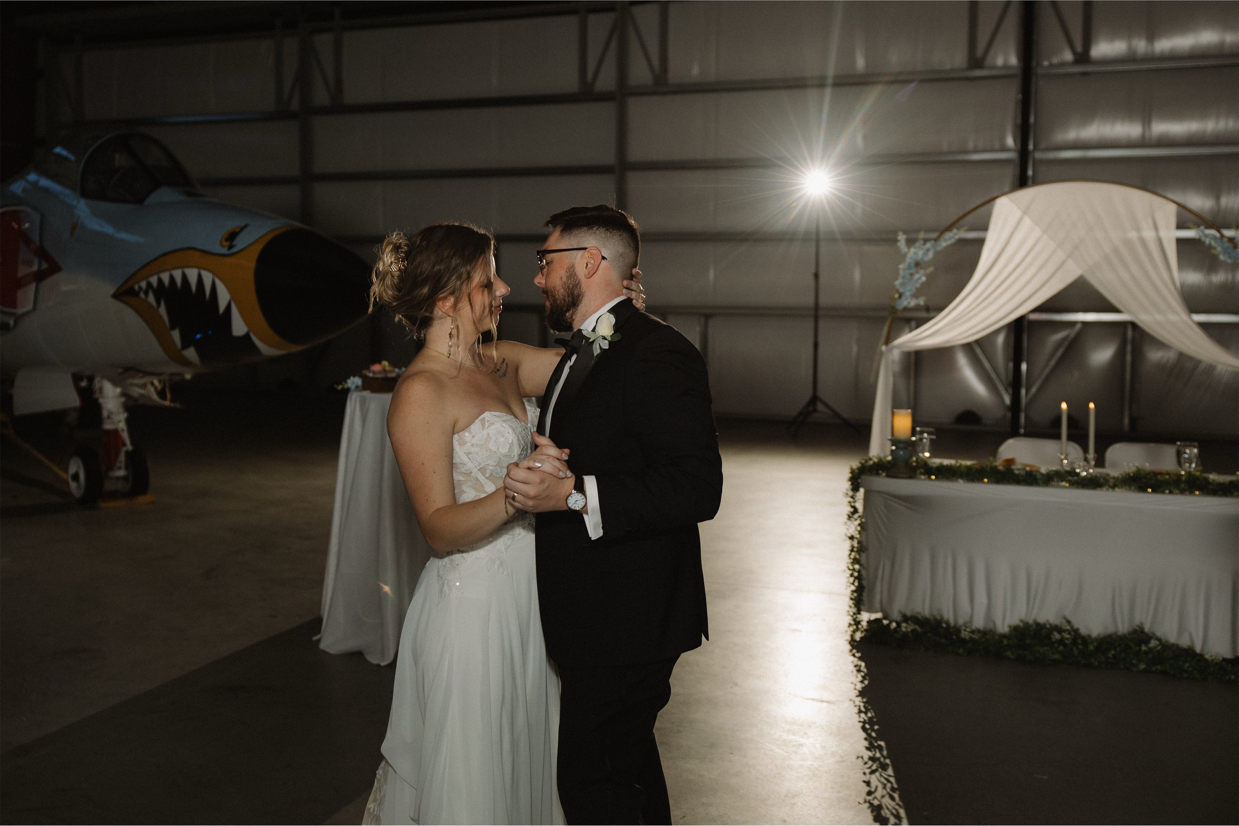A bride and groom dancing closely at their wedding reception inside a hangar, with a jet aircraft in the background and a decorated table nearby.