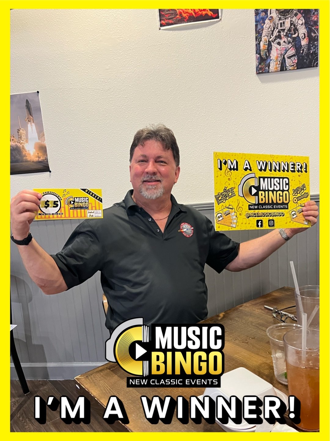 A man sitting at a restaurant table holding winning bingo tickets for Music Bingo event, smiling, with signs reading 'I'm a Winner!' and 'Music Bingo, New Classic Events'.