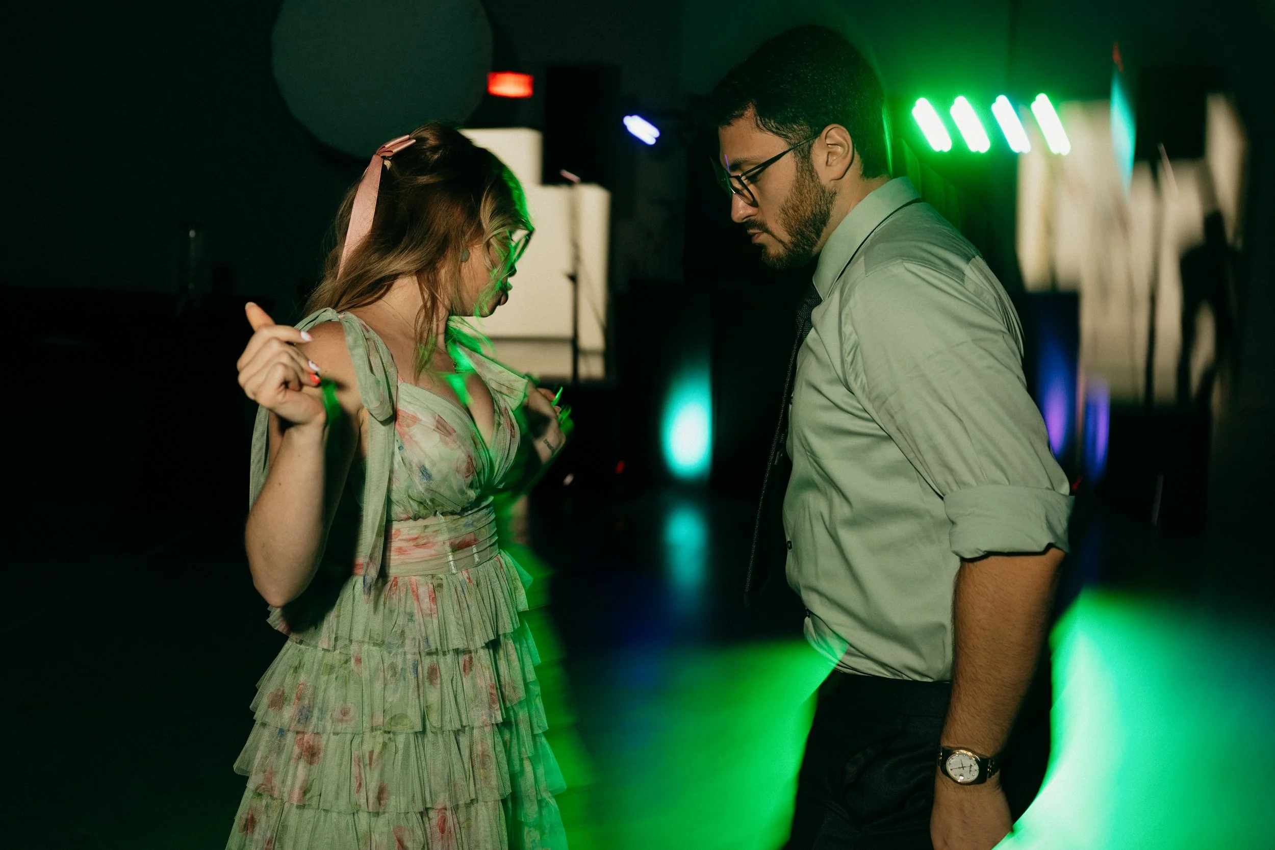 A man and woman are dancing closely at a party or nightclub, with colorful neon lights illuminating the dark background.