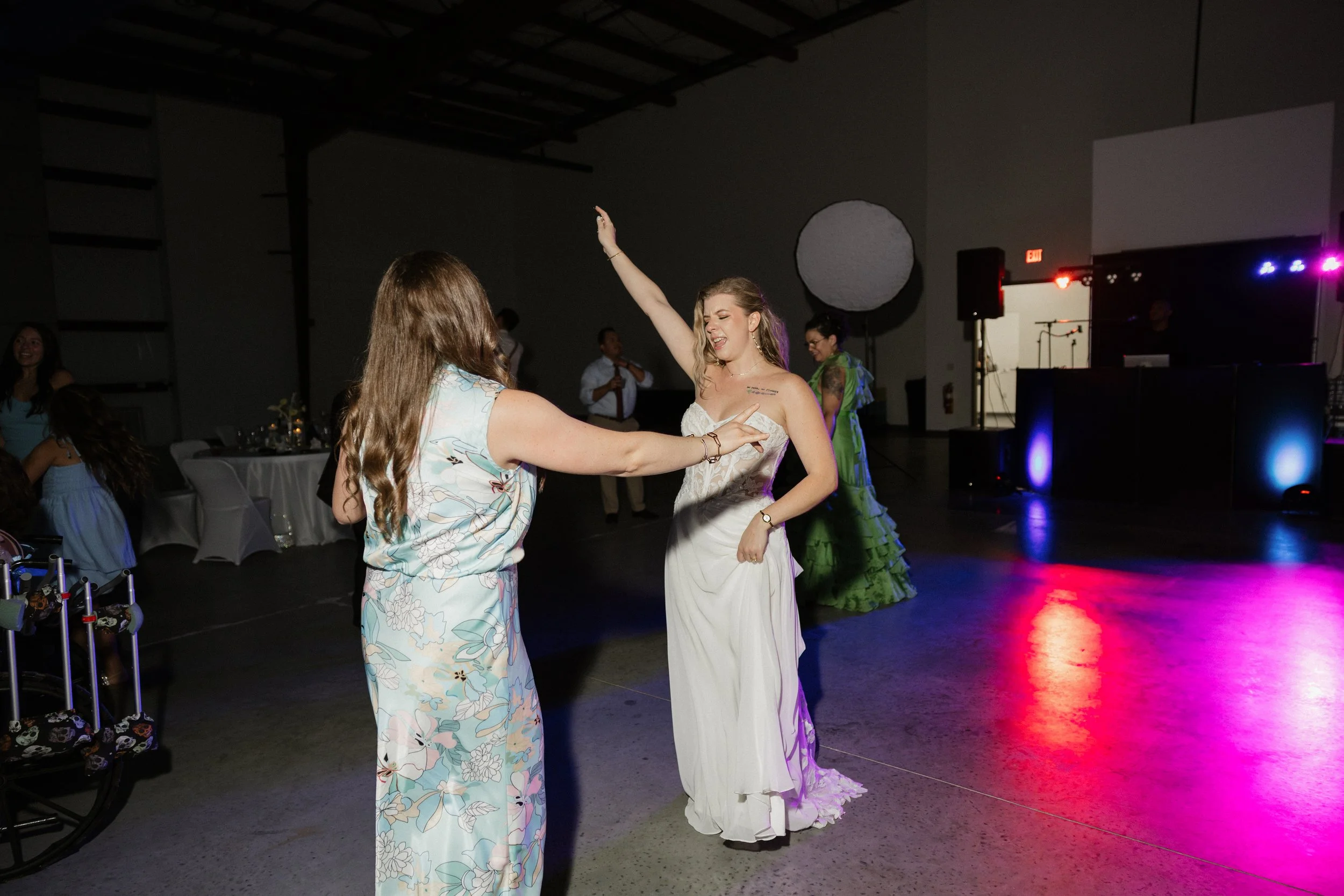 Two women dancing at a celebration, one in a white wedding dress and the other in a floral dress, with a DJ booth and colorful lighting in the background.