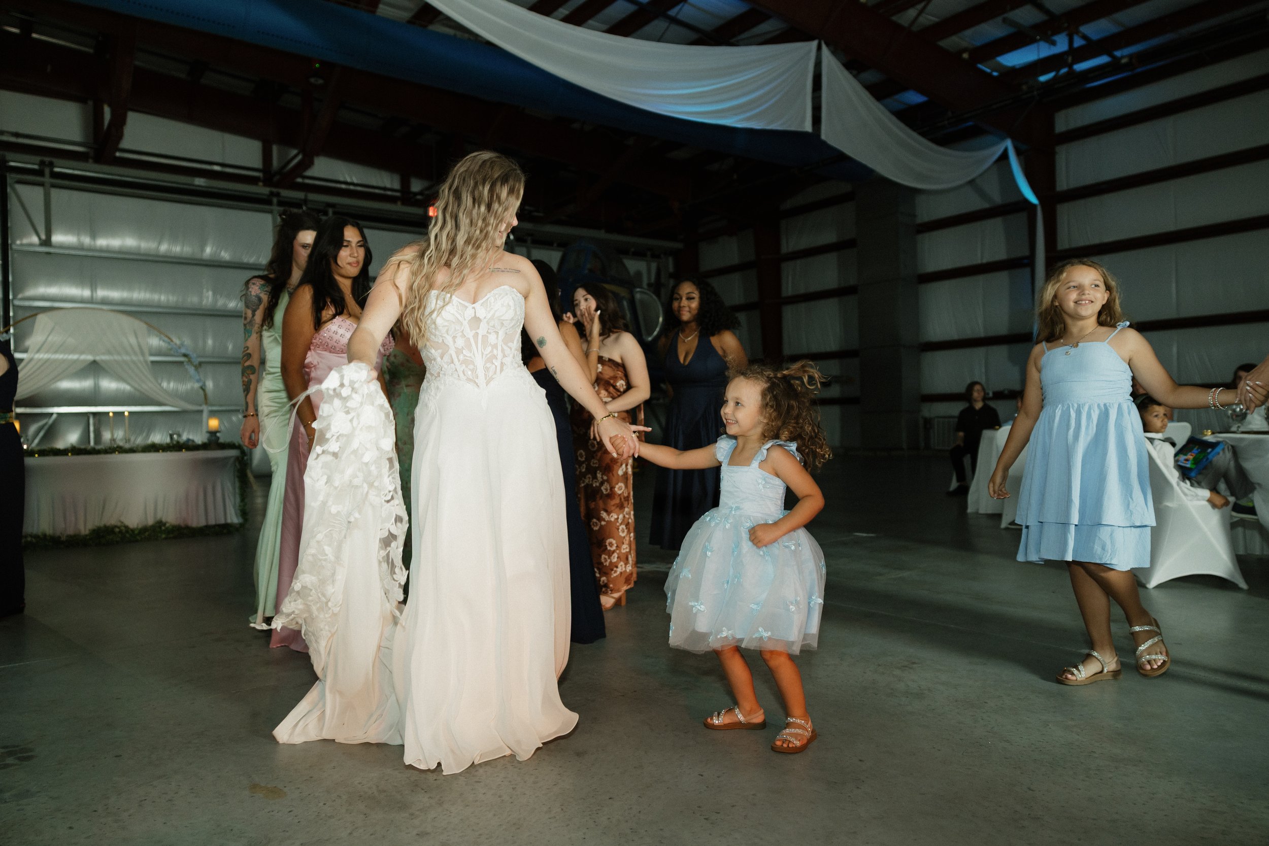 Women and girls dancing at a wedding reception in a spacious indoor venue with industrial-style architecture and high ceilings. The bride in a white gown is holding hands with a young girl in a light blue dress. Other women in dresses stand nearby, w