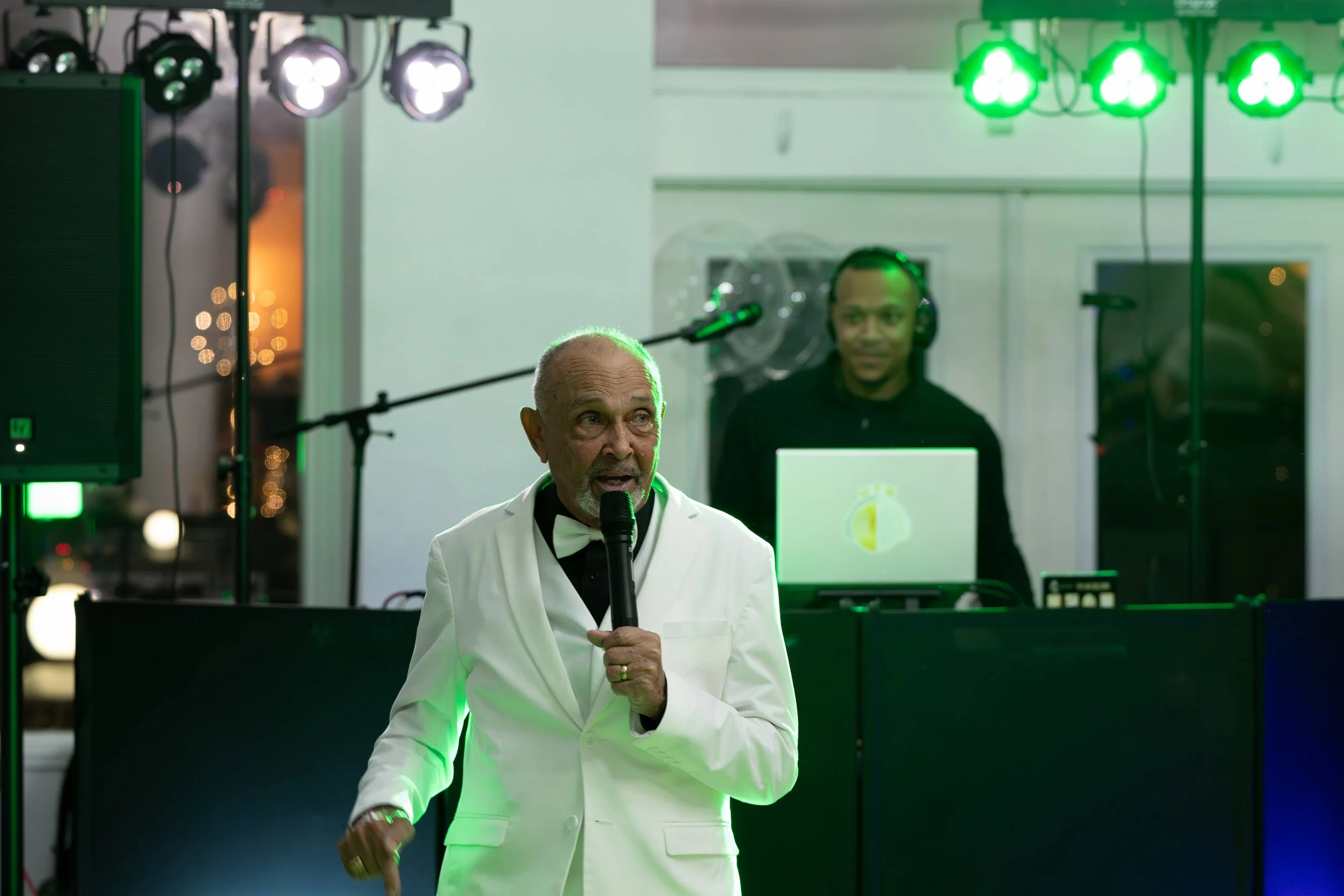 An elderly man in a white tuxedo jacket and black bow tie, holding a microphone, appears to be speaking or singing at an event. Behind him, a DJ is working at a booth with green lighting and equipment.