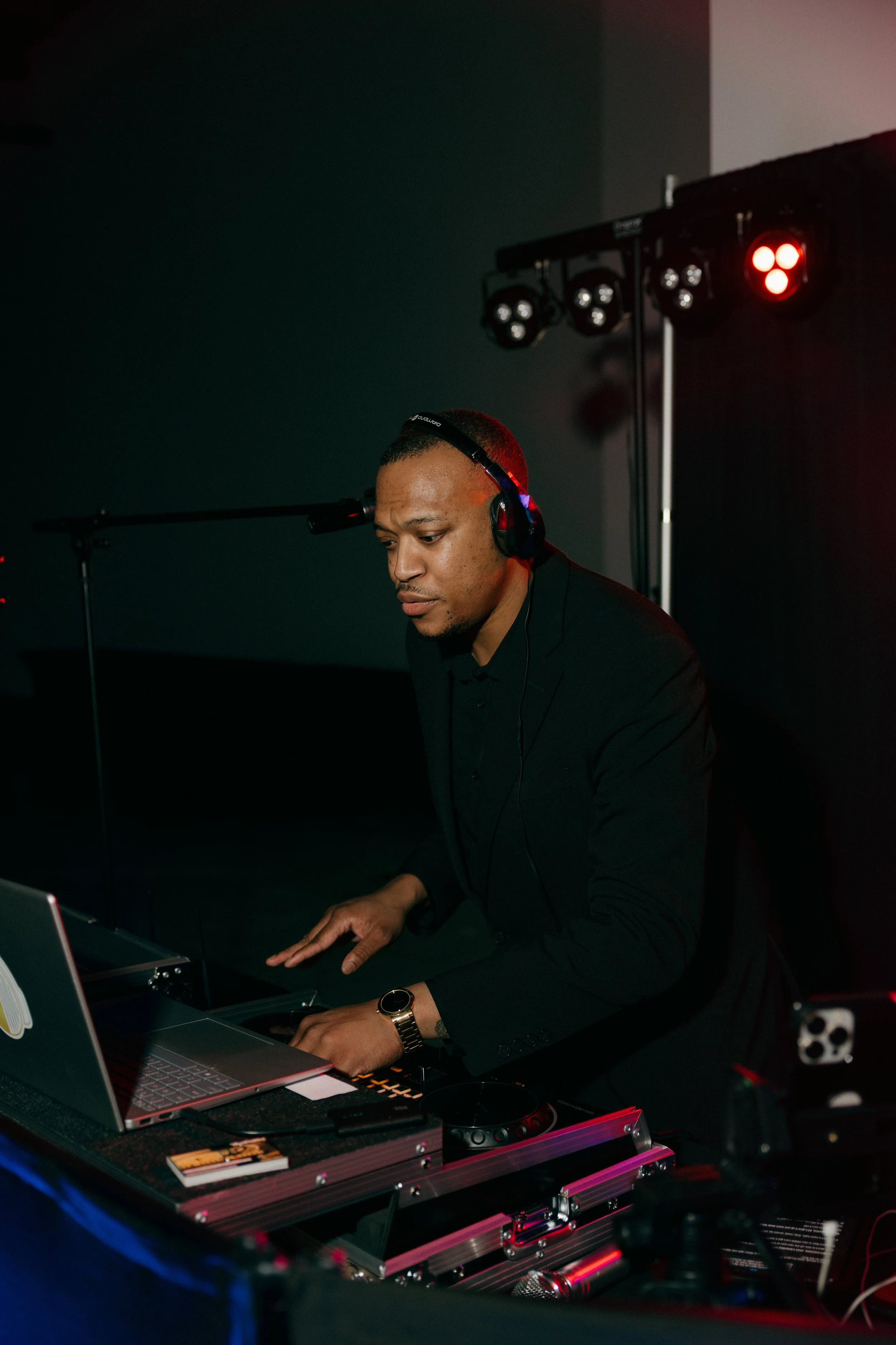 A man in a black suit and gold watch is DJing at a dark venue, wearing headphones, with DJ equipment and a laptop on a black table, surrounded by stage lighting.