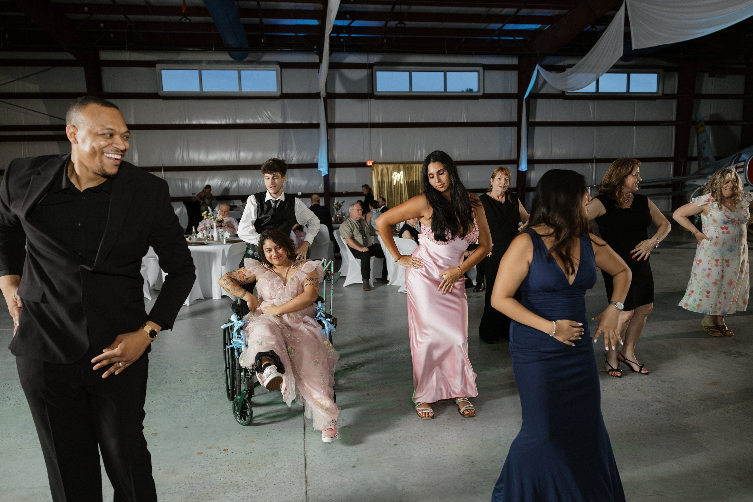 People dancing at an indoor event, including a man in a black suit, a woman in a pink dress, and a woman in a blue dress, with seated guests and a small airplane in the background.