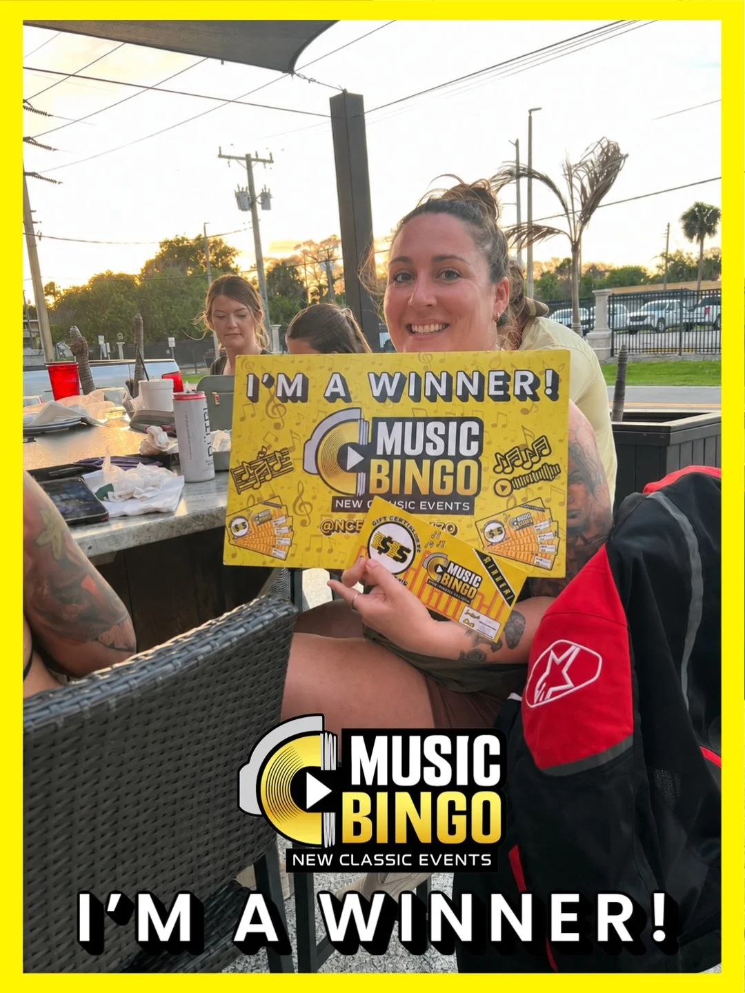 A woman holding a yellow Music Bingo certificate at an outdoor dining area, smiling with people and tables around, during sunset.