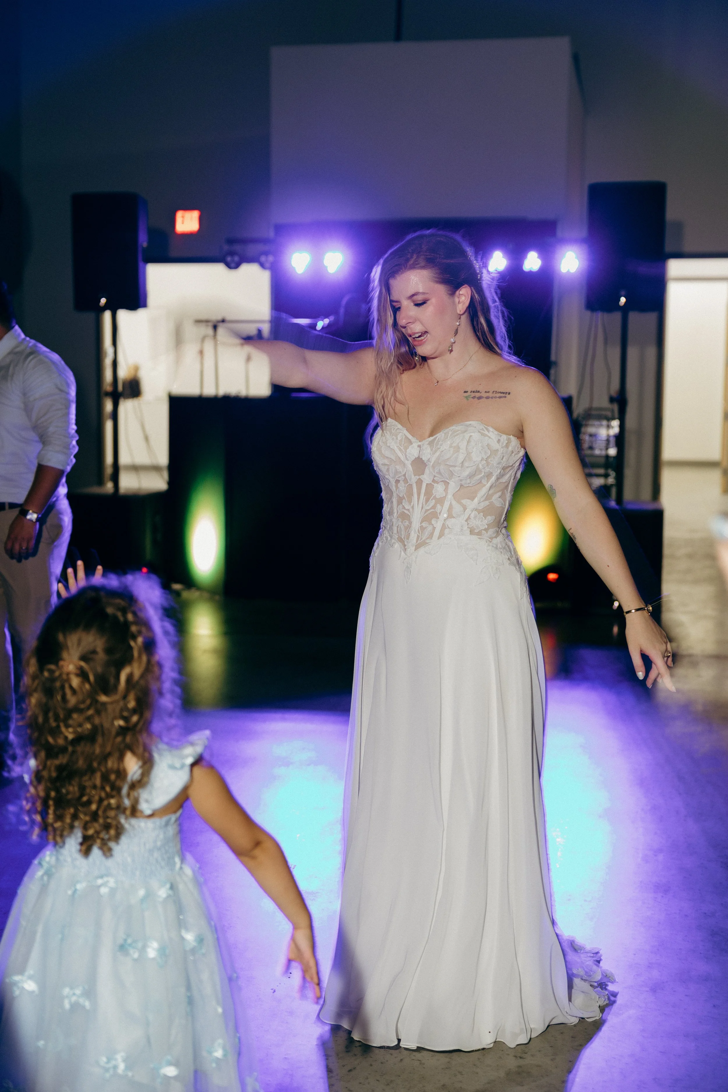 A woman in a strapless white wedding dress dances at a reception, with purple and green lighting and a DJ in the background. She has long hair and tattoos on her chest and arm, and is surrounded by children in white dresses.