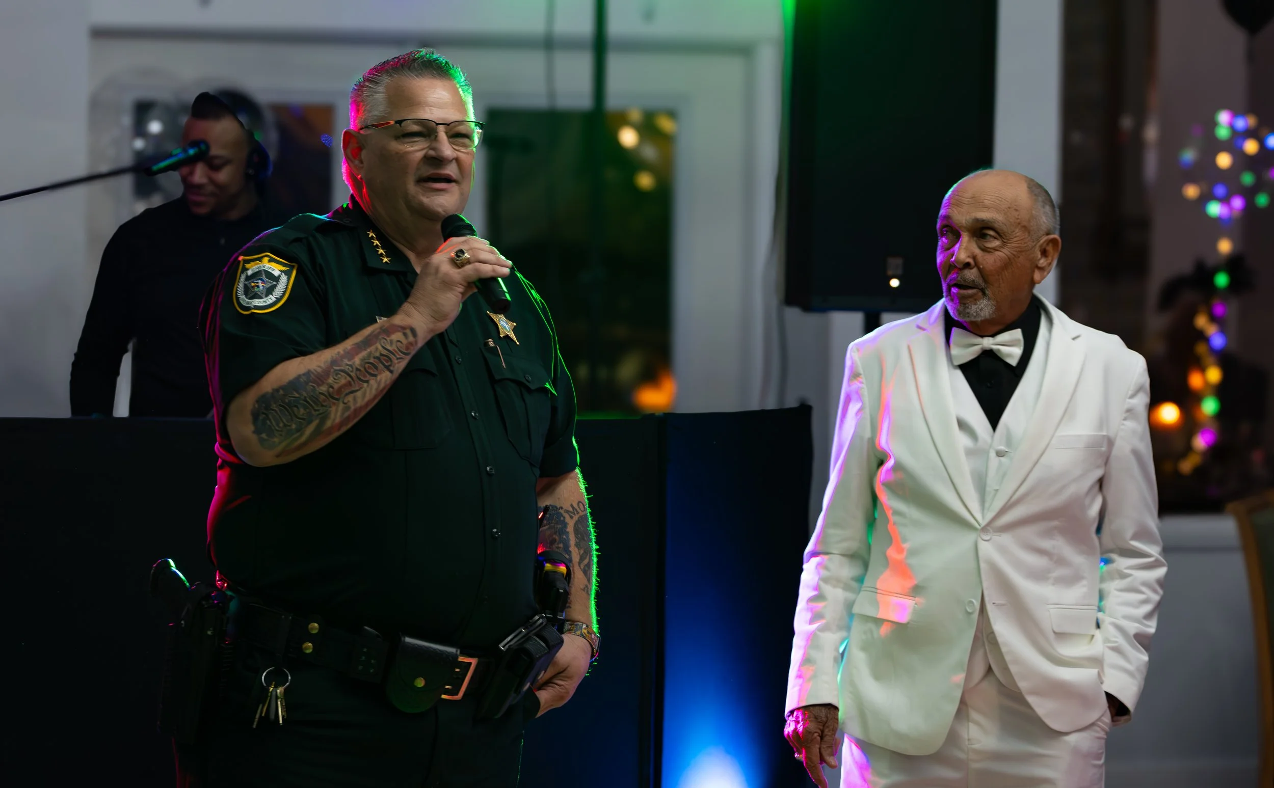 A police officer in uniform speaking into a microphone and a man in a white tuxedo listening at a decorated indoor event.
