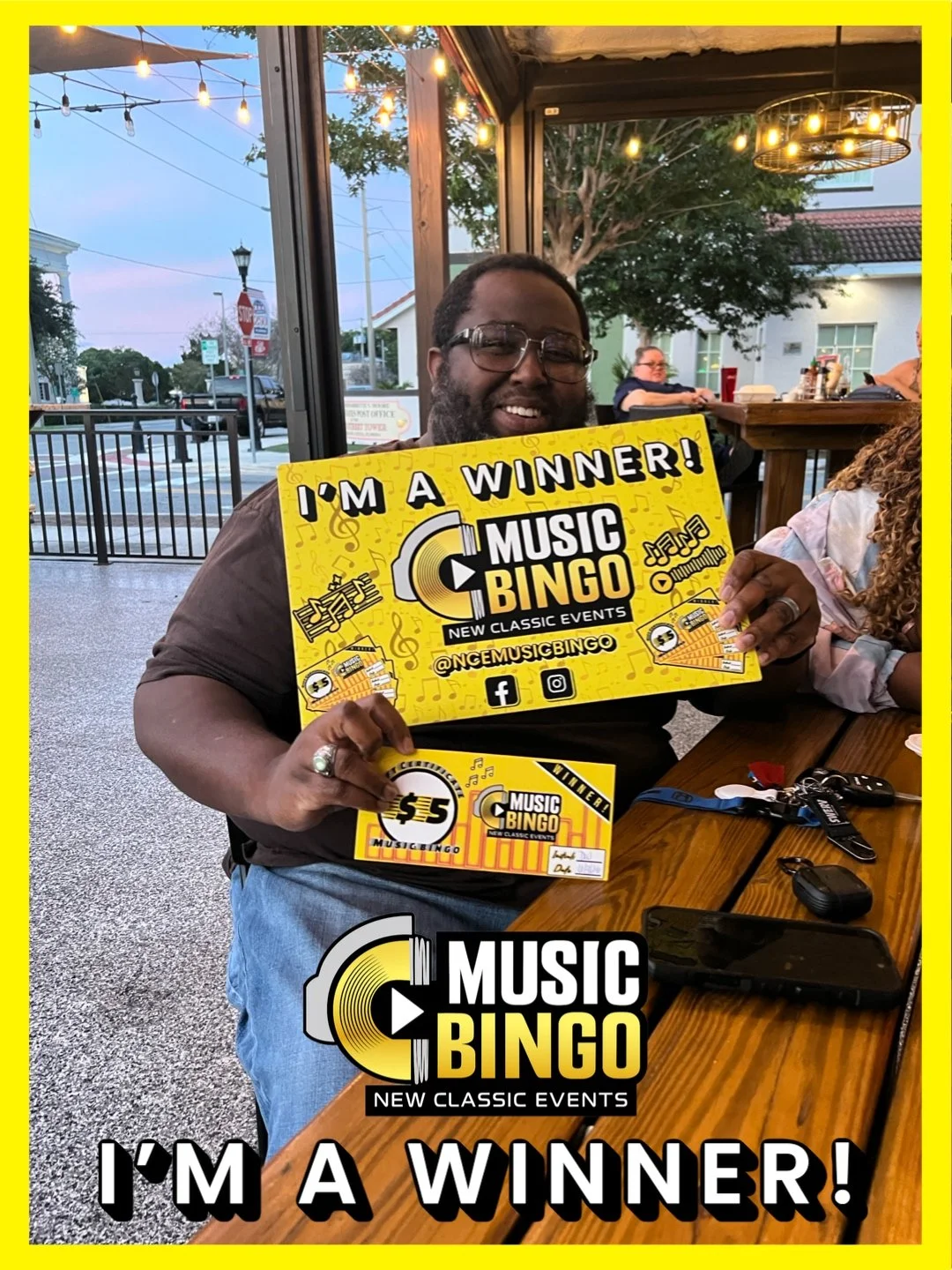 A man sitting at a table inside a restaurant, holding a yellow bingo card and a large sign that reads 'I'm a winner! Music Bingo, New Classic Events.' He is smiling, wearing glasses, and has a beard. The background shows outdoor street view and other