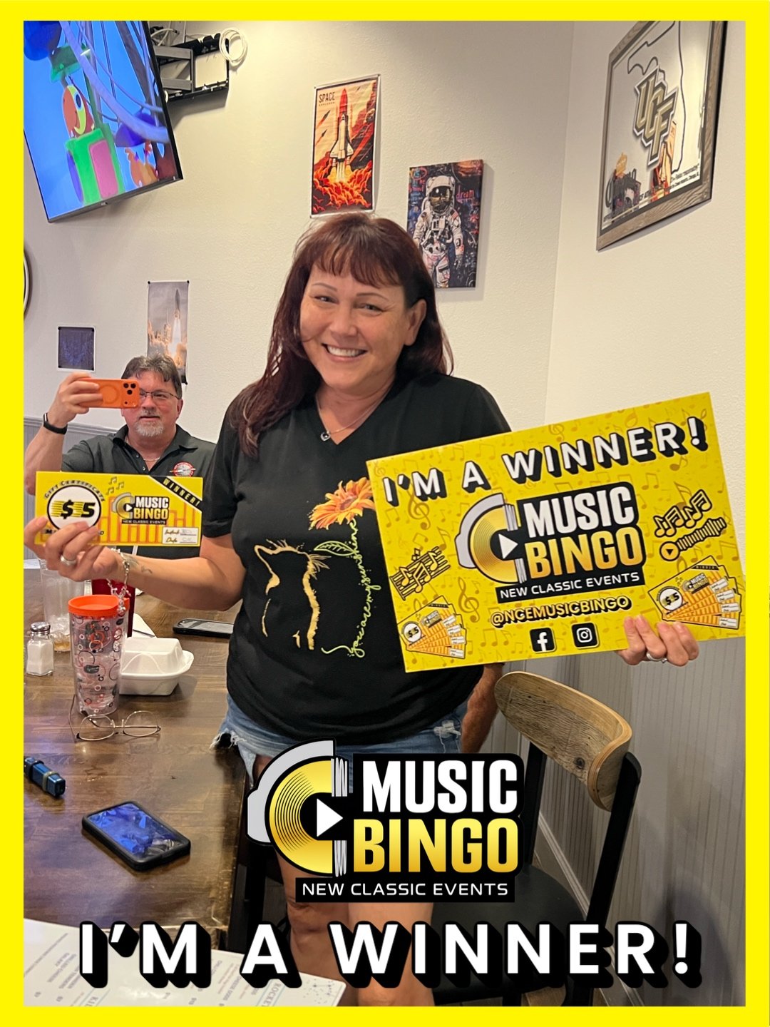 A woman smiling and holding a yellow sign that reads 'I'm a winner! MUSIC BINGO' with social media information, standing at a table in a casual setting, while a man in the background takes a photo with his phone. The room has posters and artwork on t