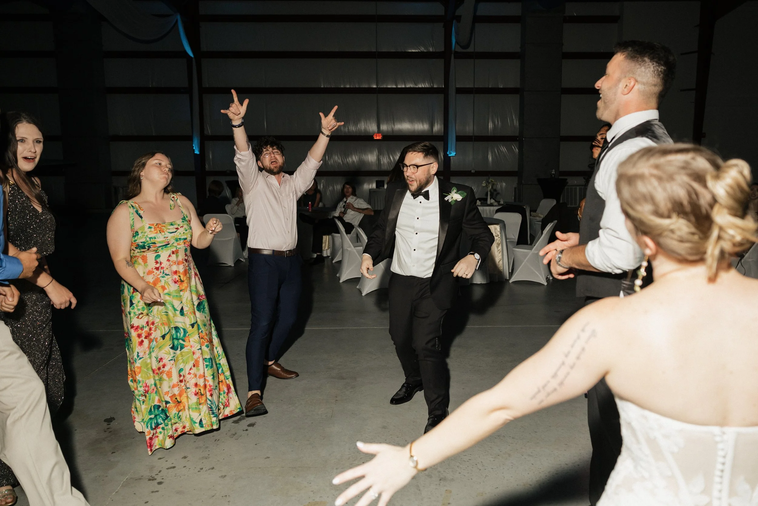 People dancing and socializing at a formal event, possibly a wedding reception, in a large hall with dark walls and sofas.