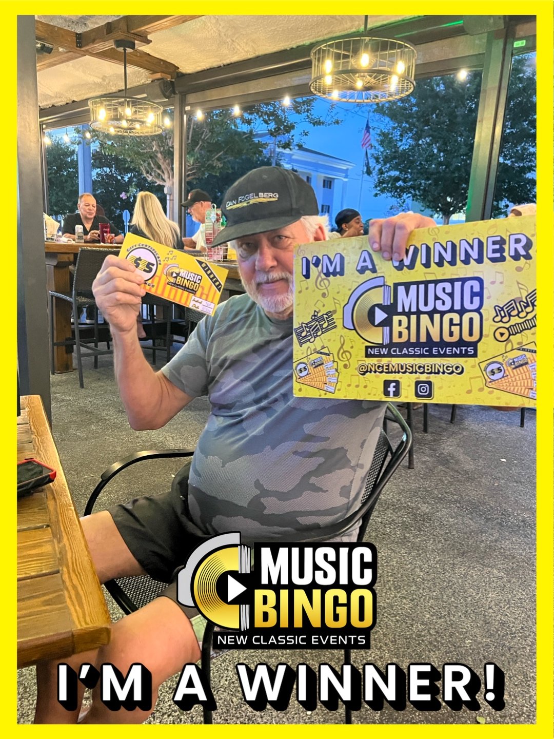 A man holding a yellow sign that says "I'm a winner Music Bingo" with musical notes and graphics, sitting at a table in a restaurant. He also holds a smaller bingo card. The background shows other people dining and decorative lighting.