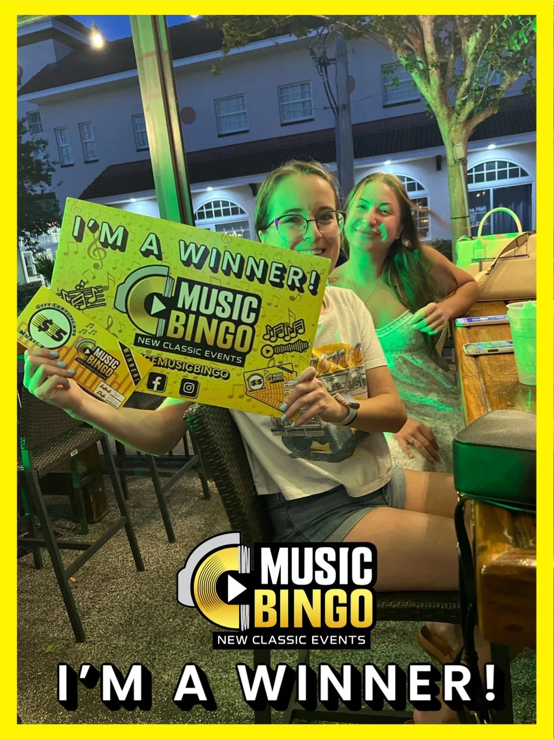 A young woman holding a yellow sign that reads "I'm a Winner!" and promoting Music Bingo at an outdoor venue during evening. She is smiling, wearing glasses and a white graphic T-shirt, with another woman smiling in the background. The scene is illum