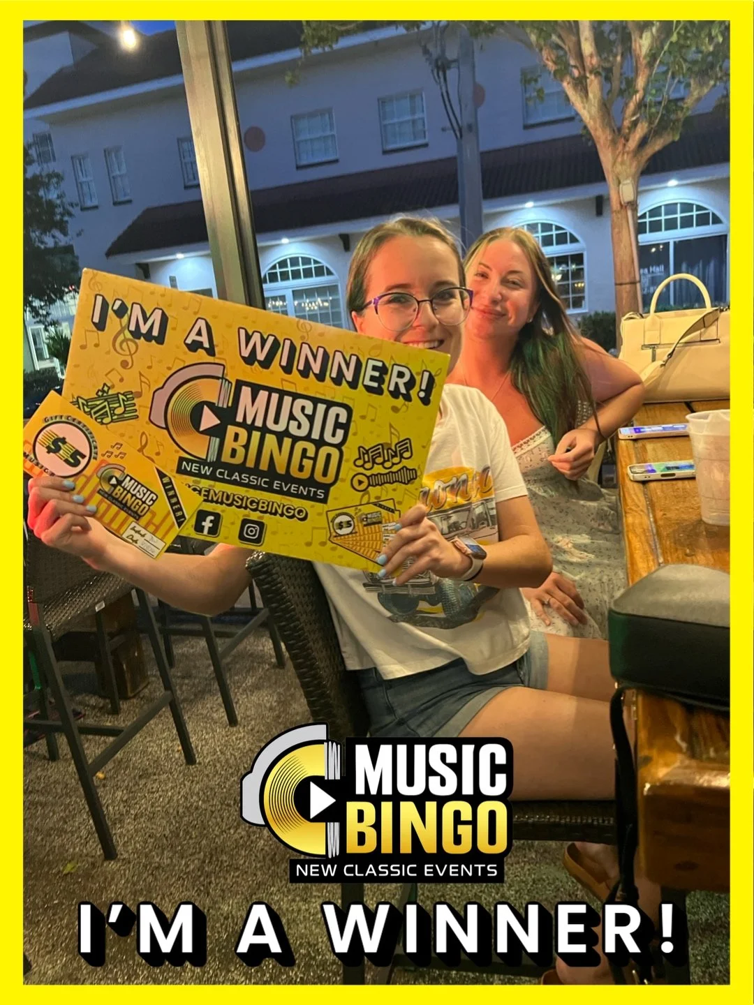 Two women sitting at a restaurant table, one holding a yellow Music Bingo sign with the words 'I'm a Winner!' and the Music Bingo logo. It is evening, with outdoor lights and building in the background.