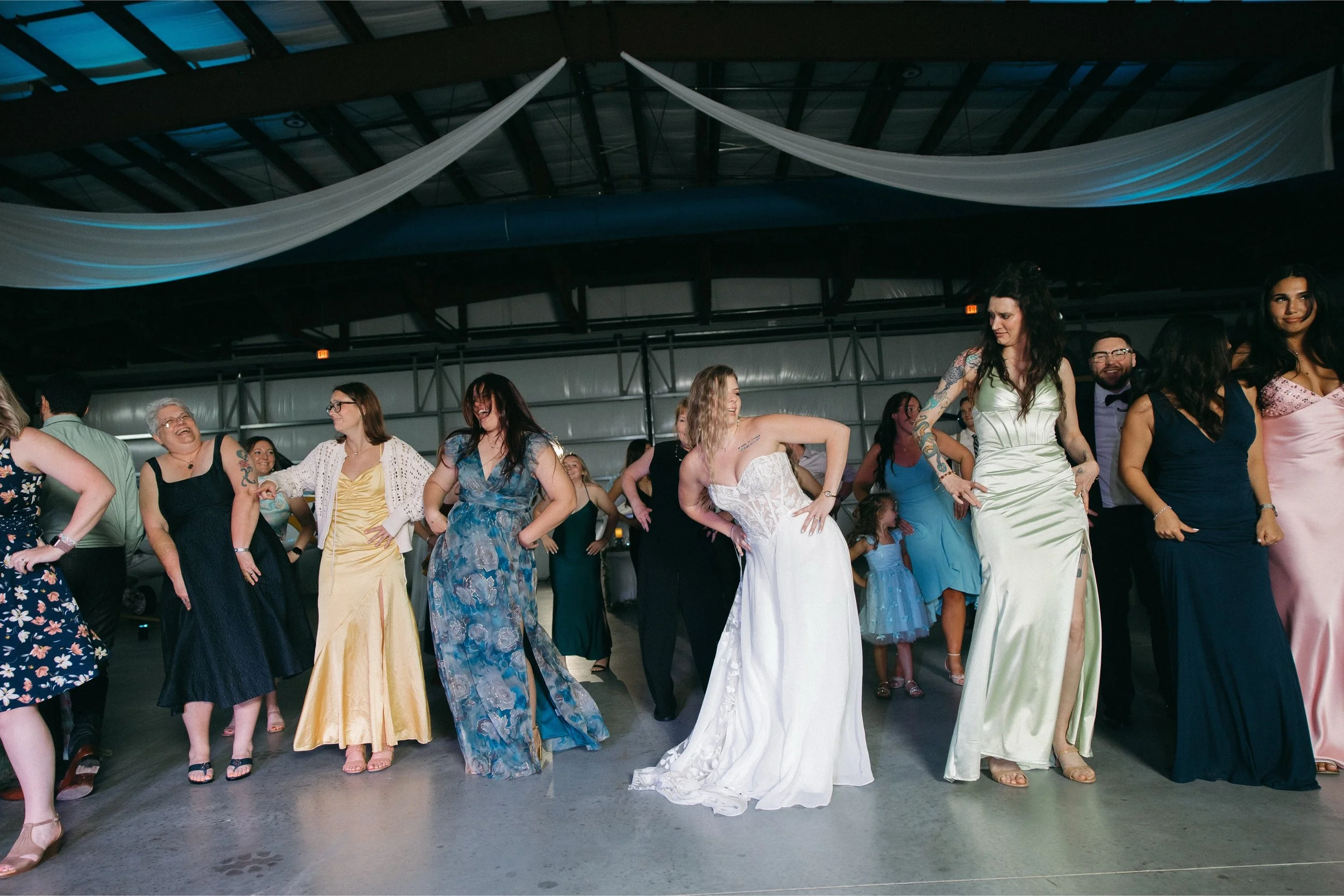 A group of people dancing at a social event, some women wearing dresses, in a large indoor space with draped fabric on the ceiling.