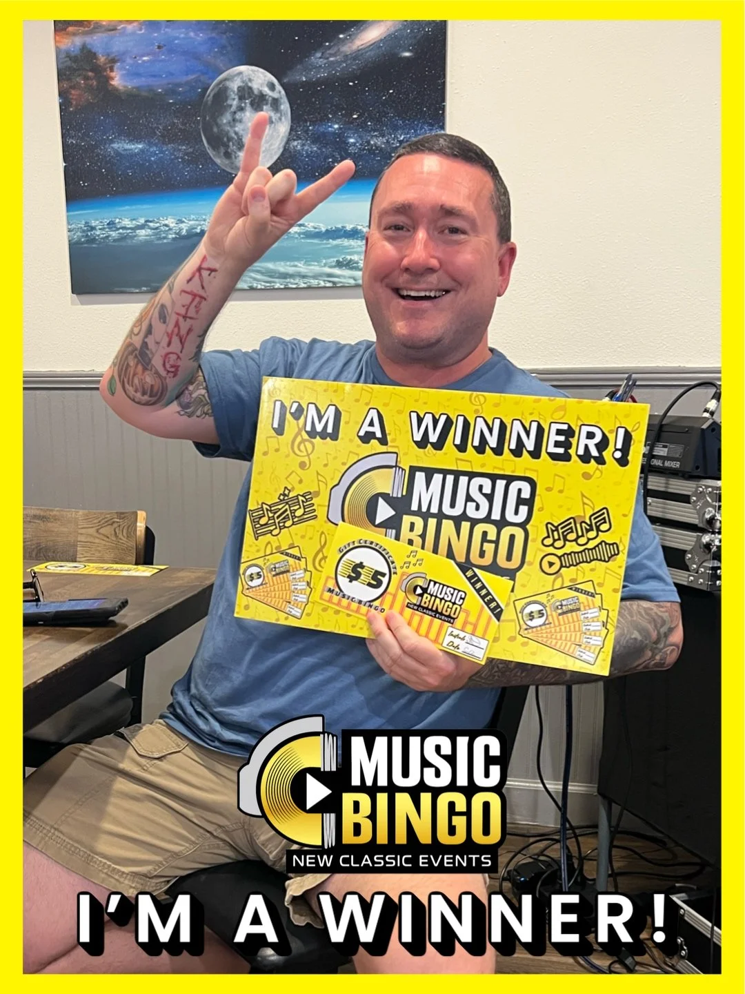 A man sitting at a table holding a yellow sign that says "I'm a Winner!" and "Music Bingo." He is smiling, making a peace sign with his right hand, and has tattoos on his left arm. Behind him, a space-themed poster depicting the moon, stars, and Eart