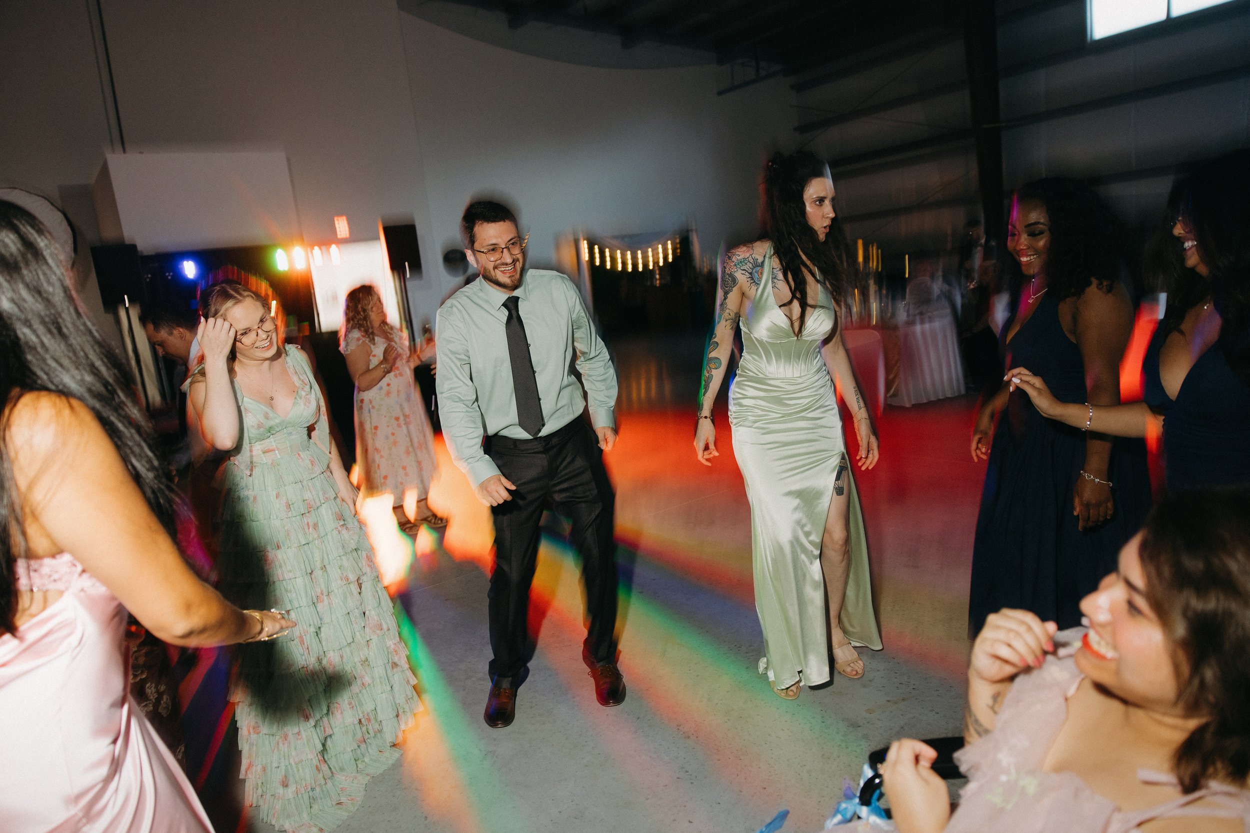 People dancing and smiling at a party or celebration in a dimly lit room with colorful lights.