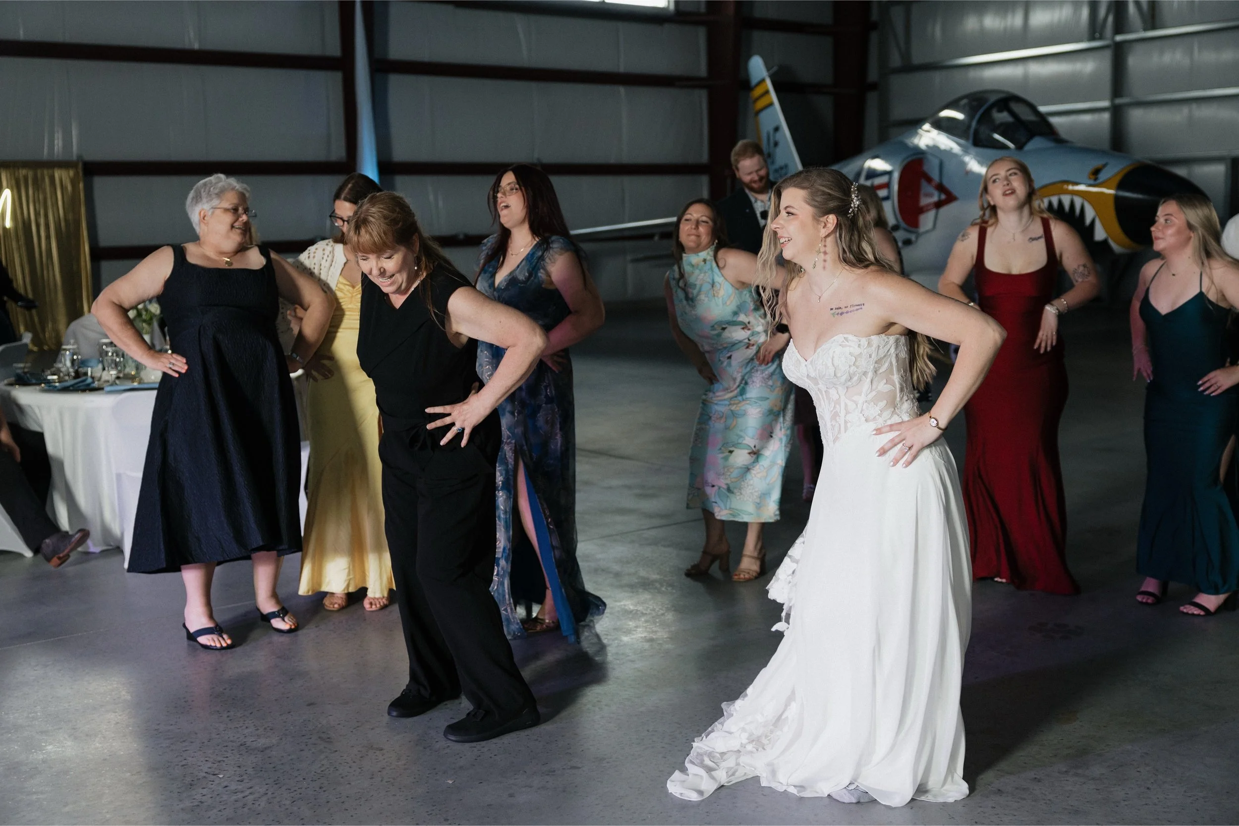 A group of women and a man dancing at a celebration in a hangar with an airplane in the background, including a bride in a white wedding dress.