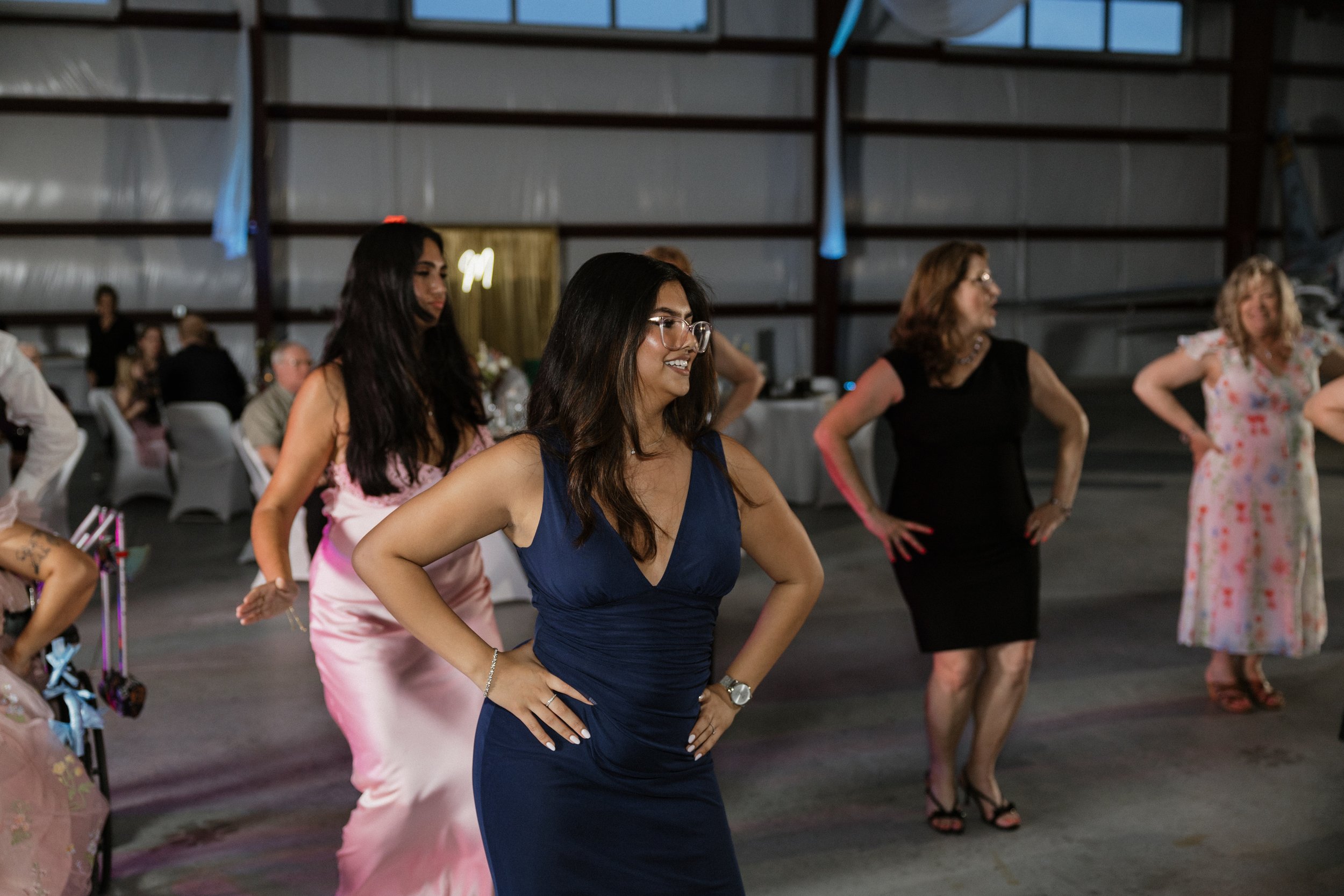 Women dancing and smiling at a social event inside a large industrial-style venue with high ceilings and windows.