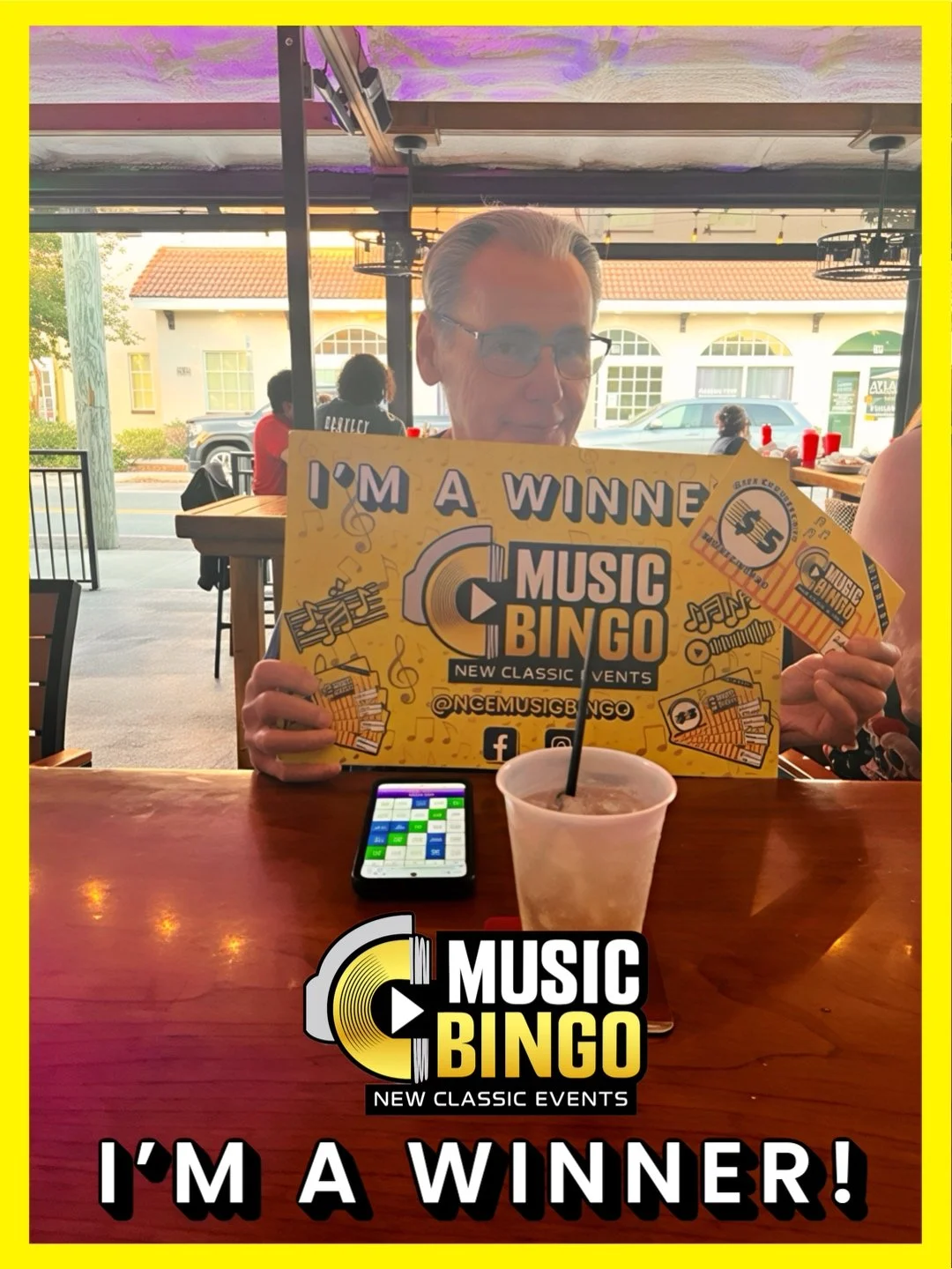 A man with glasses seated at a restaurant table holding a large promotional sign for a music bingo event that says 'I'm a Winner!'. The table has a smartphone and a drink with a straw. The background shows other patrons and a window with a street vie