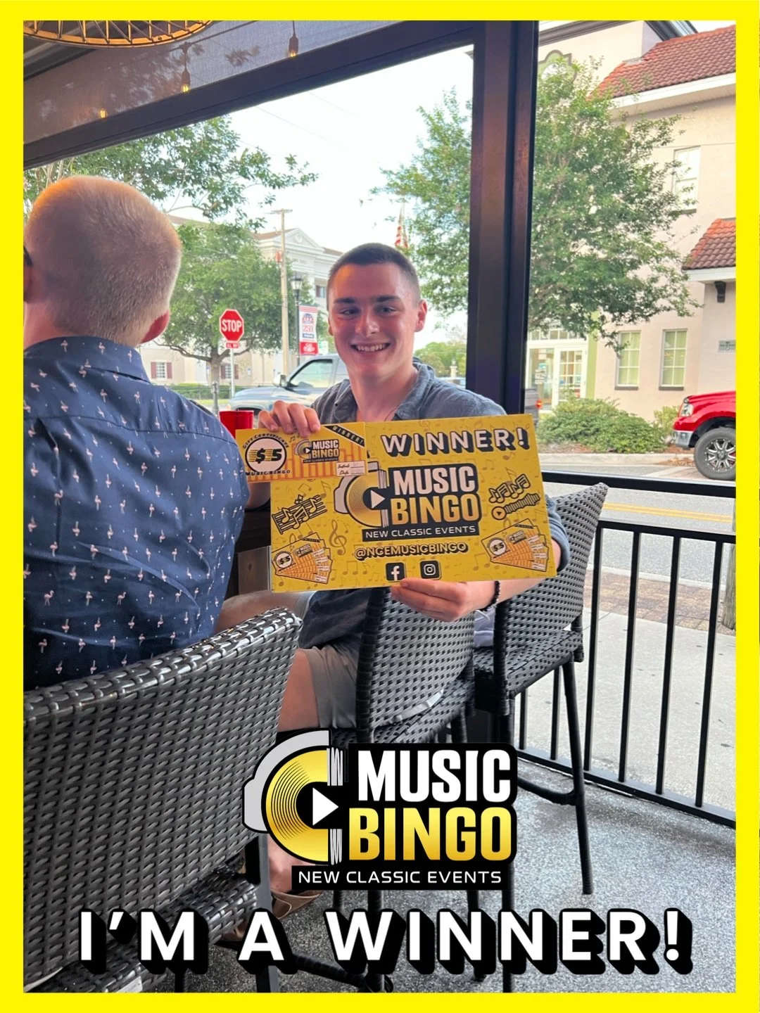 A young man with short hair smiling and holding a yellow prize certificate that says 'Winner! Music Bingo' inside a cafe, with another person sitting across from him. Outside the window, there are trees, buildings, and cars on the street.