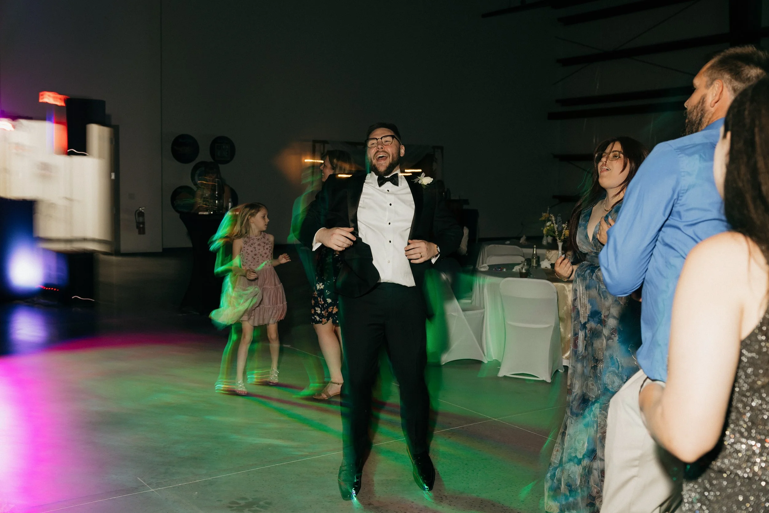 People dancing and socializing at an indoor event or party, with a man in a tuxedo in the center laughing and having fun.