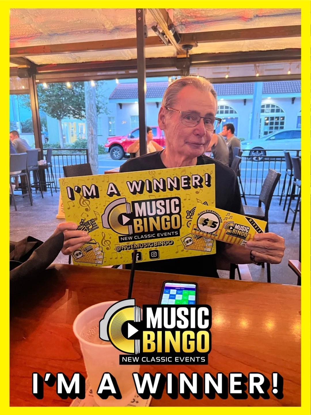 An elderly man sitting at a table indoors, holding a large yellow banner and a small printed ticket for 'Music Bingo,' with a cup on the table and people in the background.
