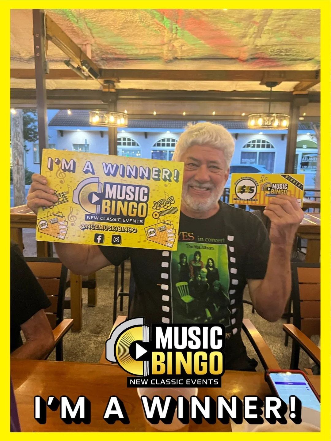 Smiling man with gray hair and beard holding a Music Bingo winner sign and a small card at a restaurant or bar with outdoor seating. He wears a black T-shirt featuring a band photo. The background shows hanging light fixtures and a building outside w