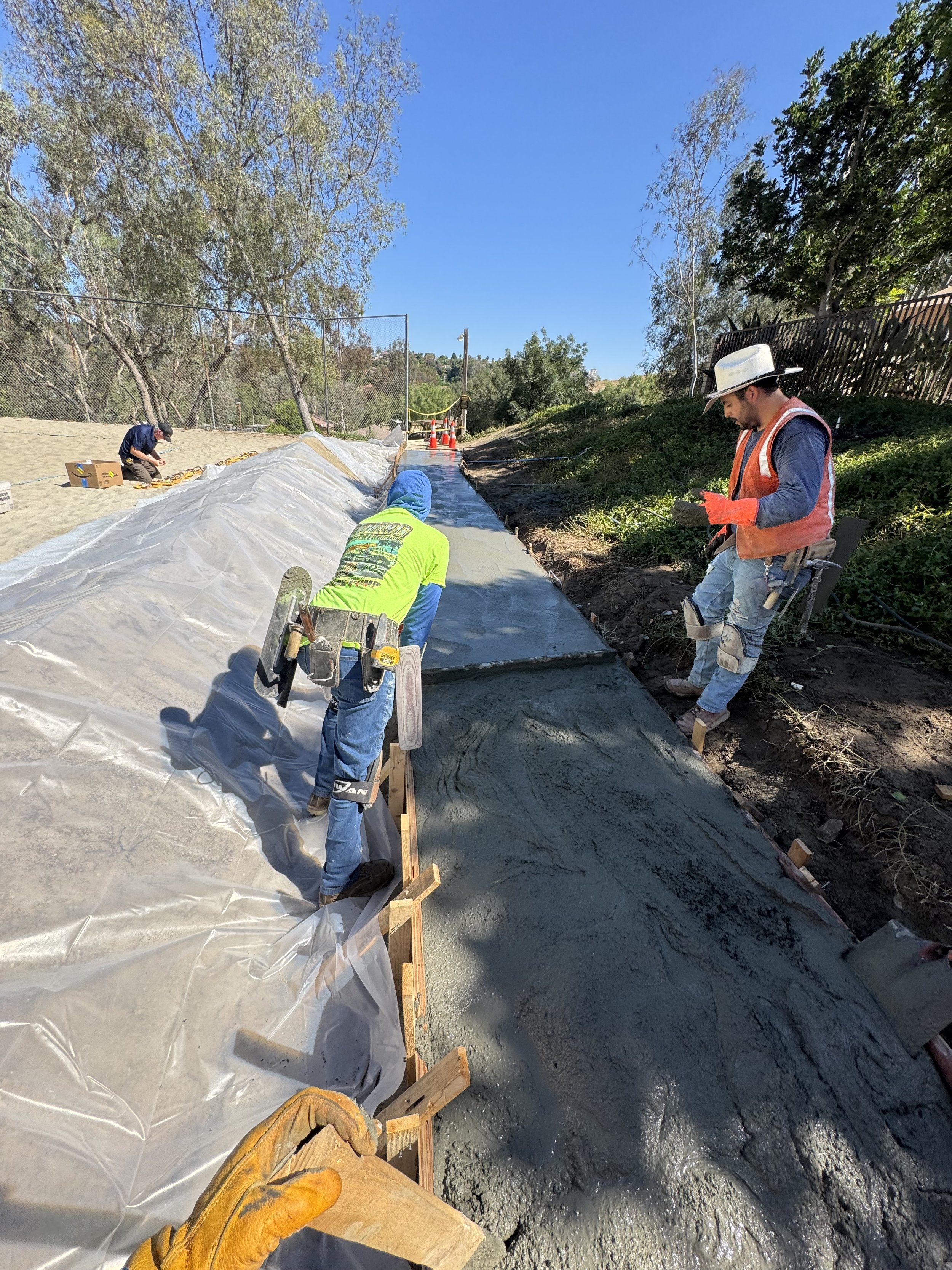 Construction workers leveling and pouring concrete on a pathway outdoors on a sunny day.