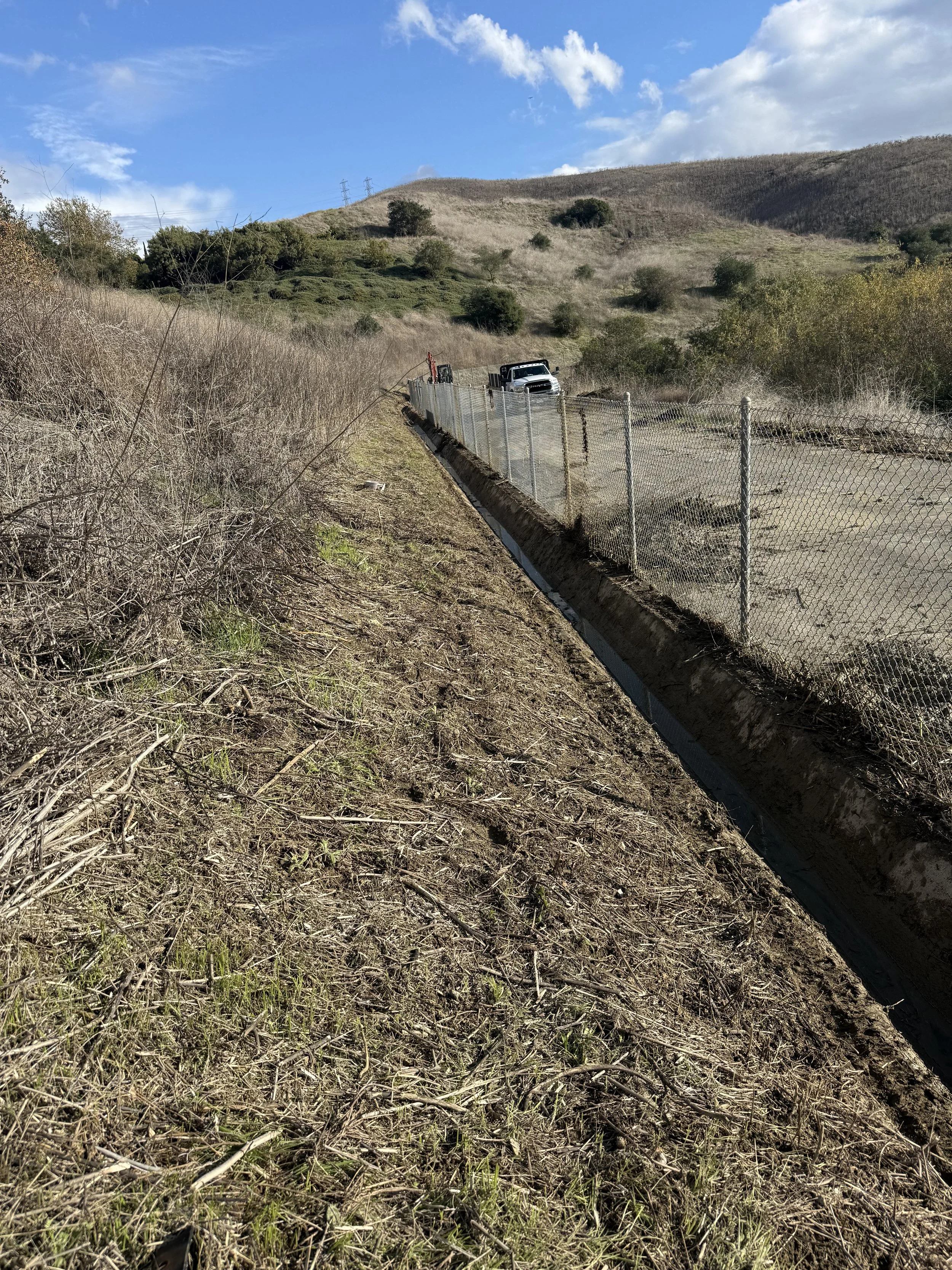 A rural hillside landscape with a dirt path next to a chain-link fence, a vehicle, and dry vegetation under a partly cloudy sky.