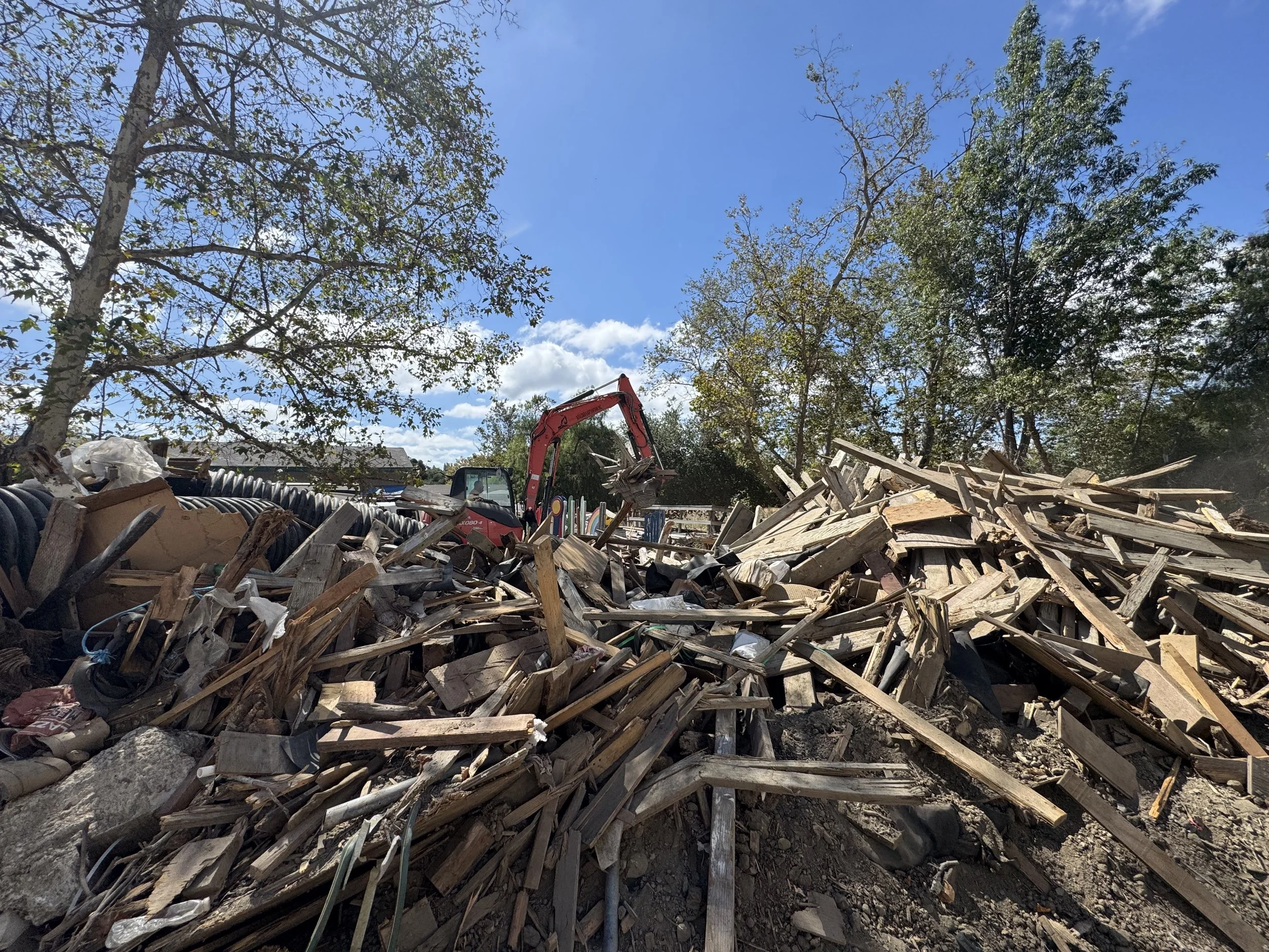 A construction site with a red excavator working on a large pile of debris and discarded wood, with trees and a blue sky with some clouds in the background.