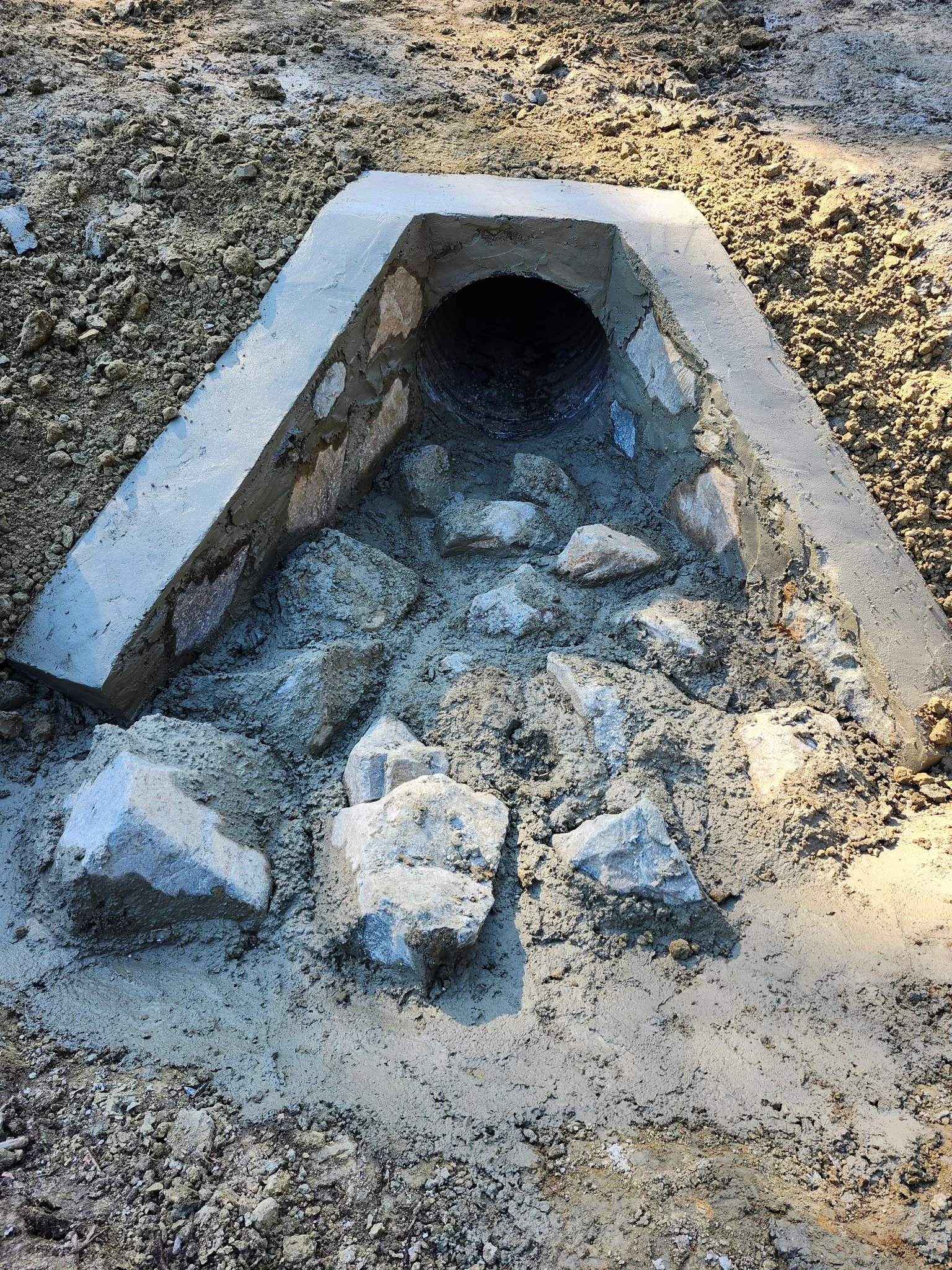 Close-up of a storm drain or culvert pipe embedded in dirt and rocks, with loose soil and rocks around it.