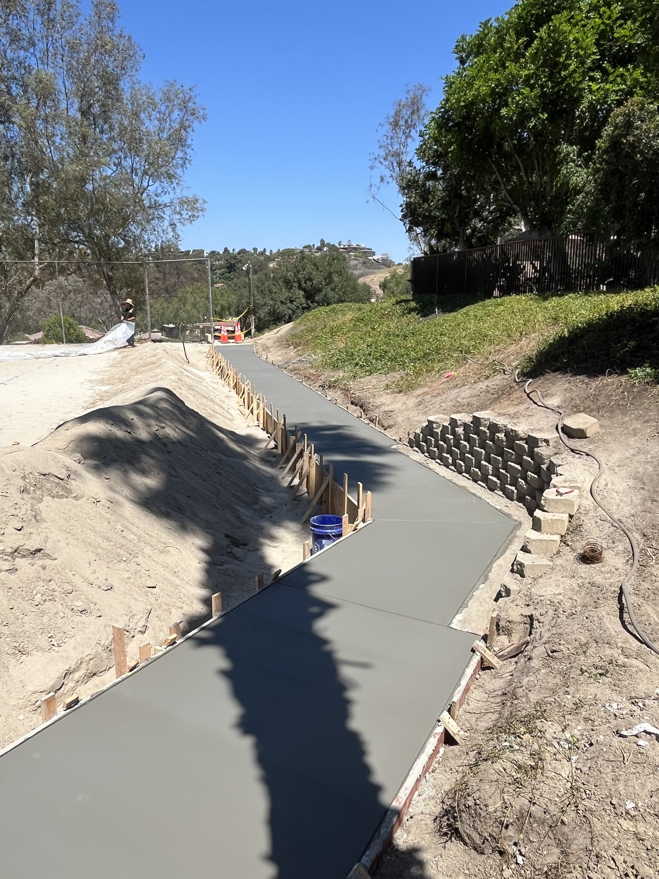 Newly poured concrete sidewalk under construction on a sunny day, with wooden forms and construction materials along the edges, and a dirt excavated area nearby.