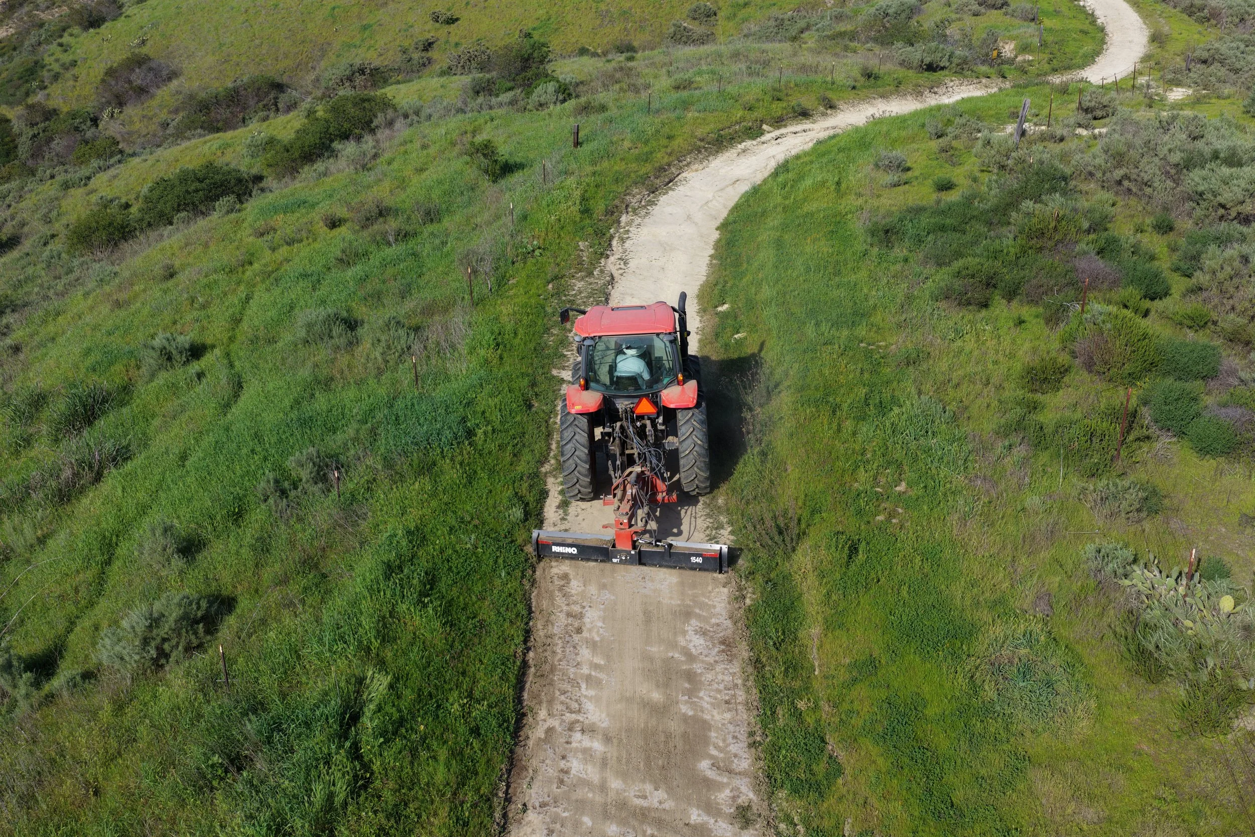 A red tractor with a wide attachment at the front leveling dirt on a winding dirt path through green, hilly terrain.