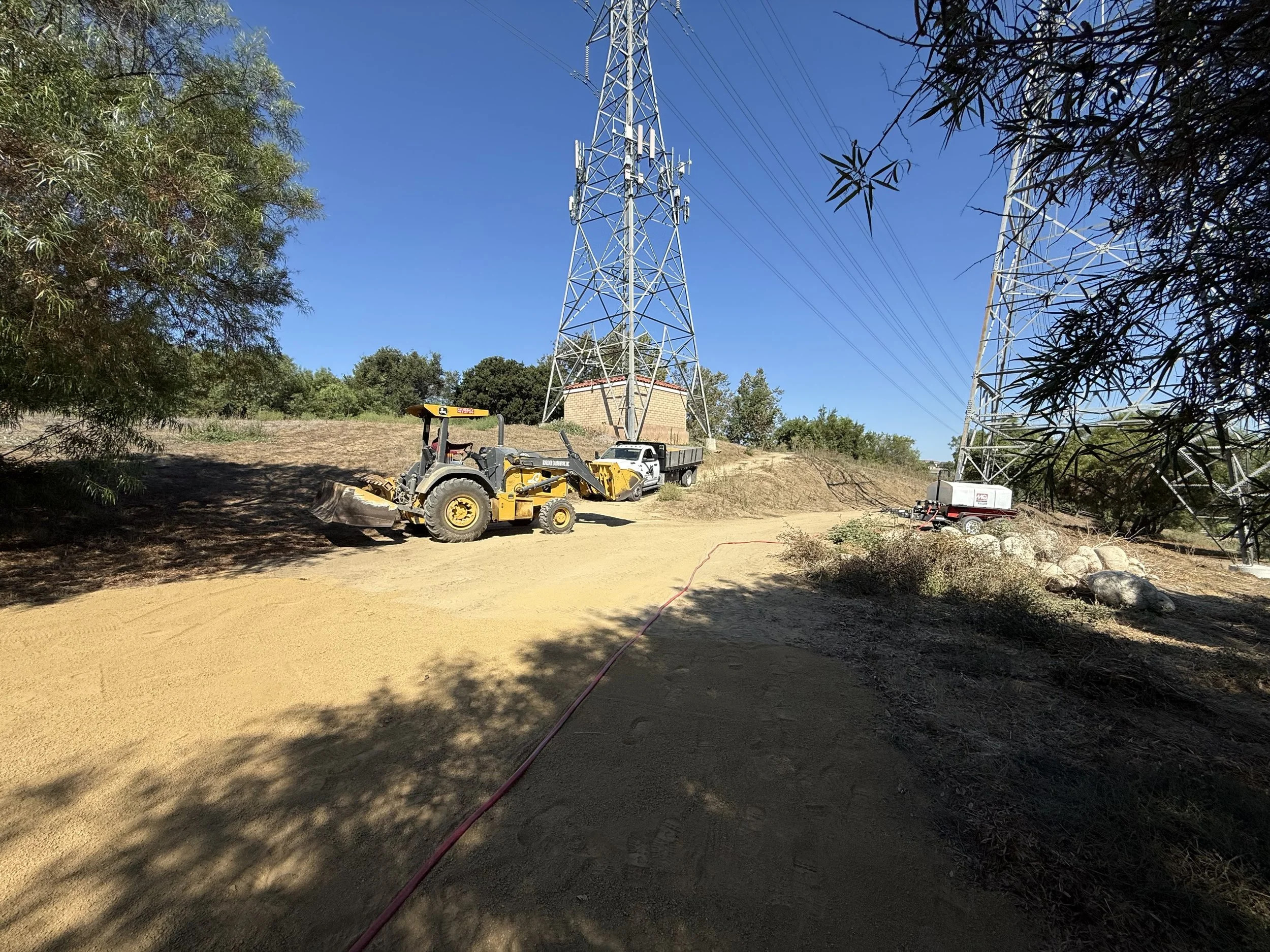 Construction site with a large electrical tower, a grader, and a utility truck on a dirt path under a clear blue sky.