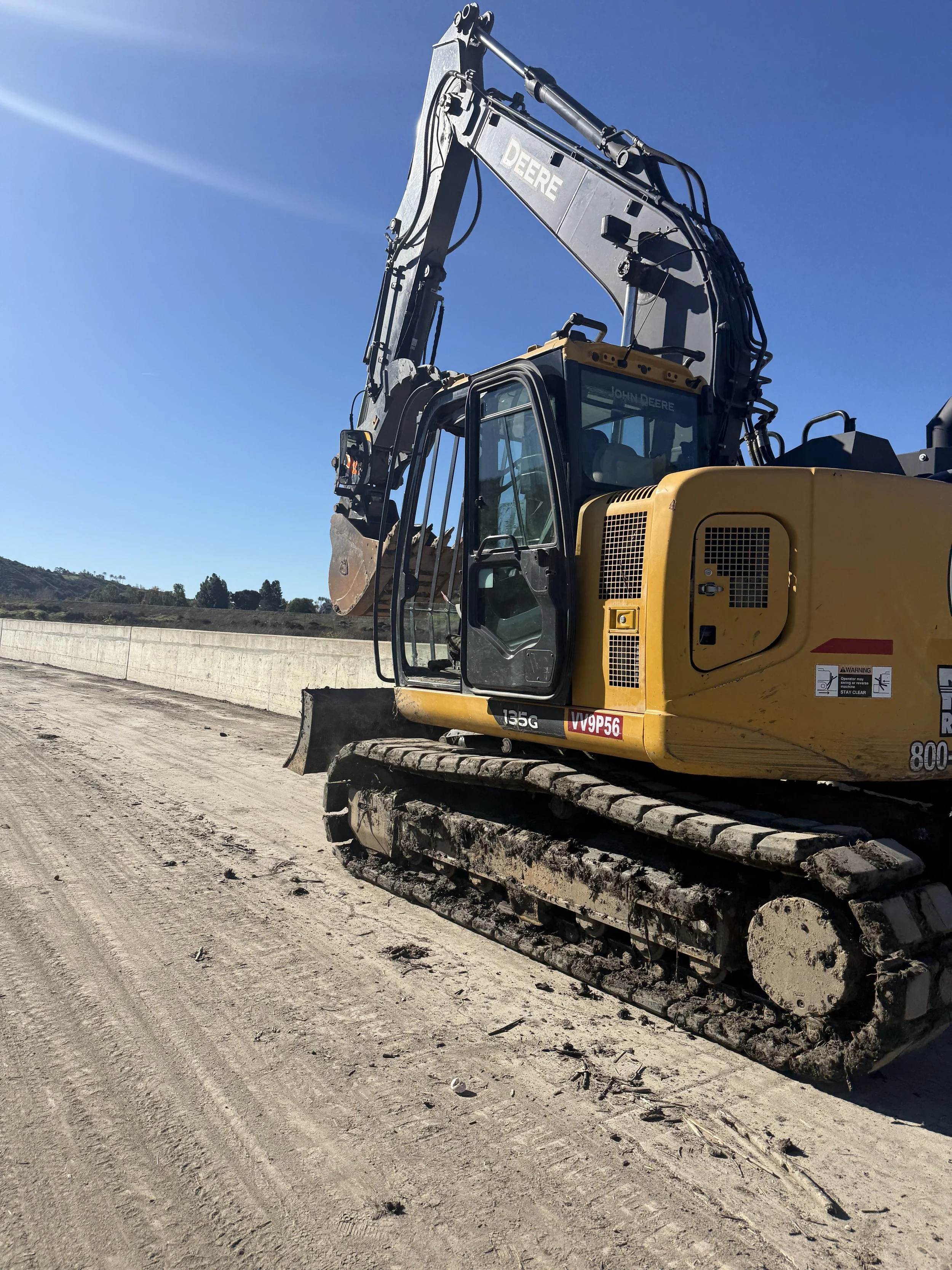 Yellow John Deere excavator on a construction site with dirt and a concrete barrier under a clear blue sky.