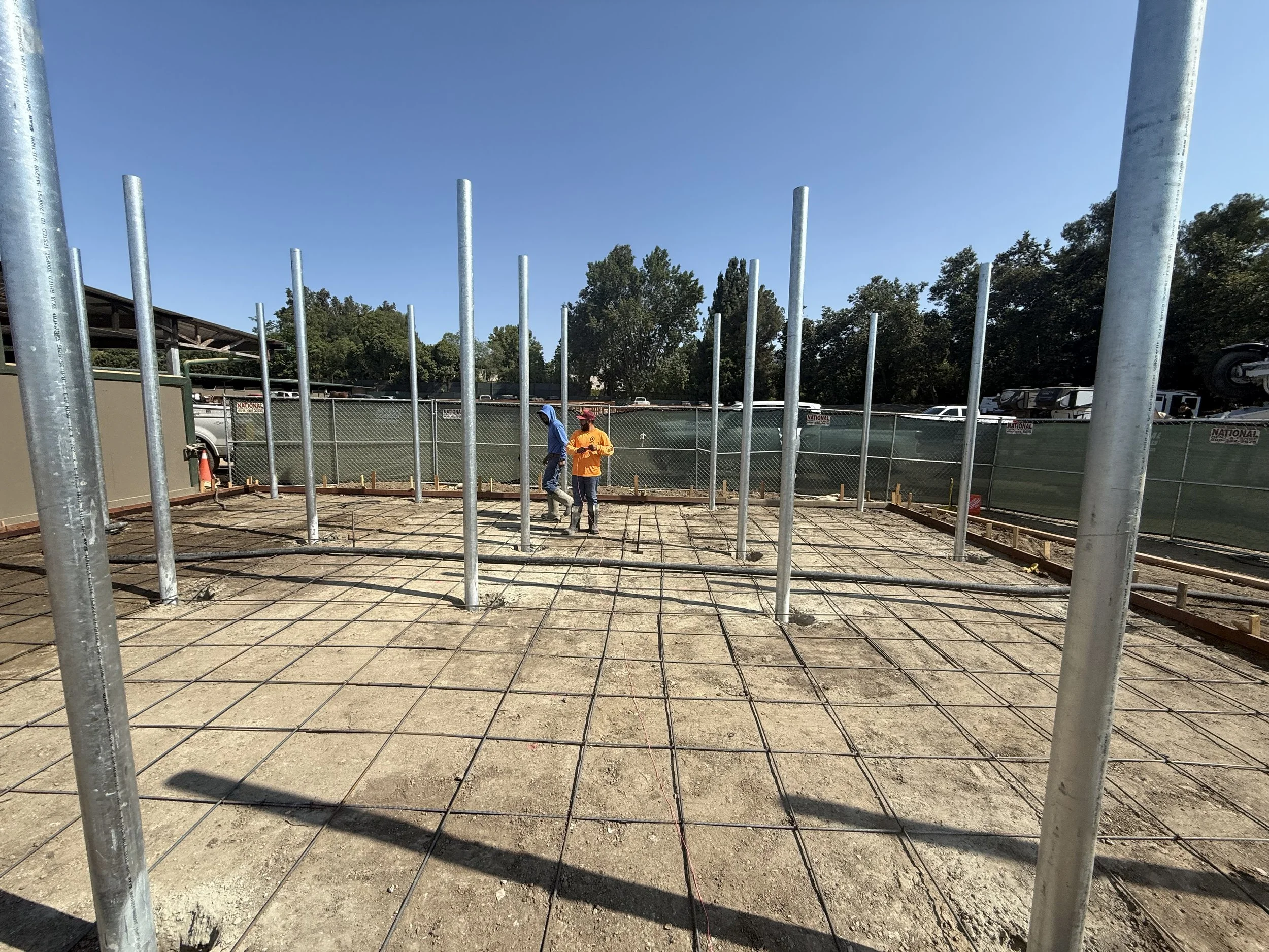Construction site with metal poles and rebar grid on dirt, two labors prepping for concrete 