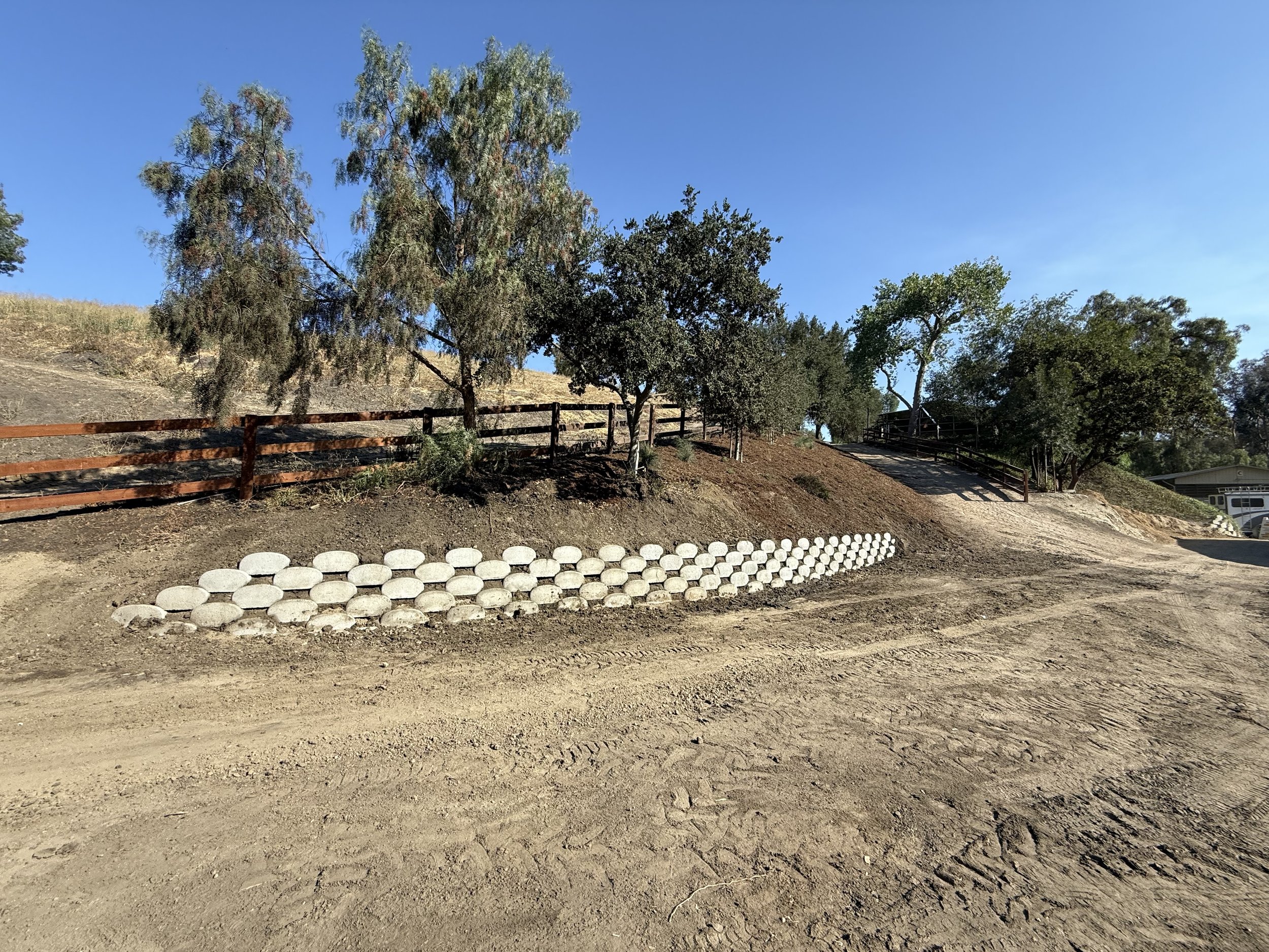 A dirt pathway with a retaining wall made of concrete rocks. A brown fence runs along the top of the slope, and there are several green trees under a clear blue sky.