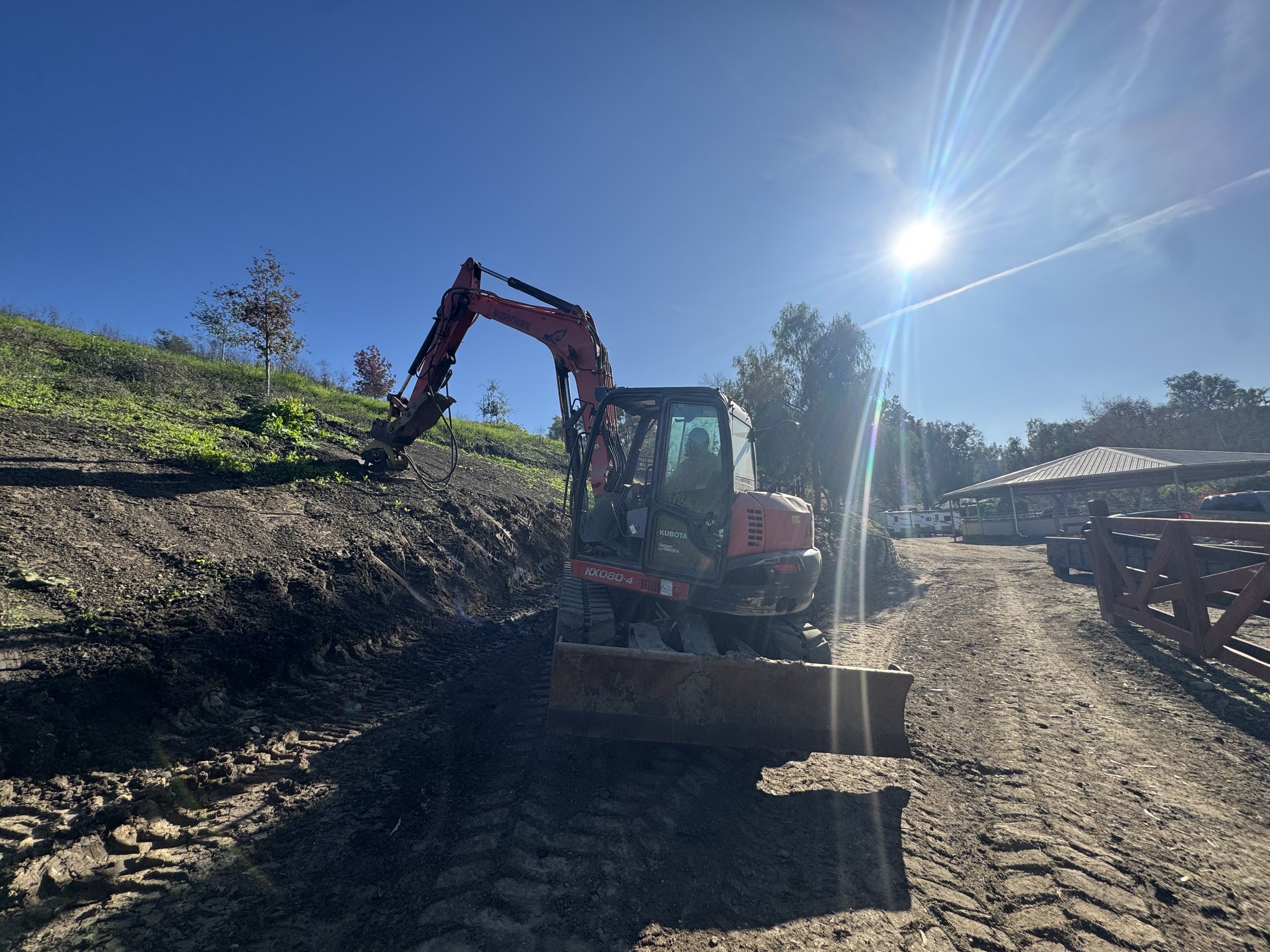 Construction site with a small excavator on a dirt pathway, hillside with small trees, and a clear sunny sky with the sun shining brightly.
