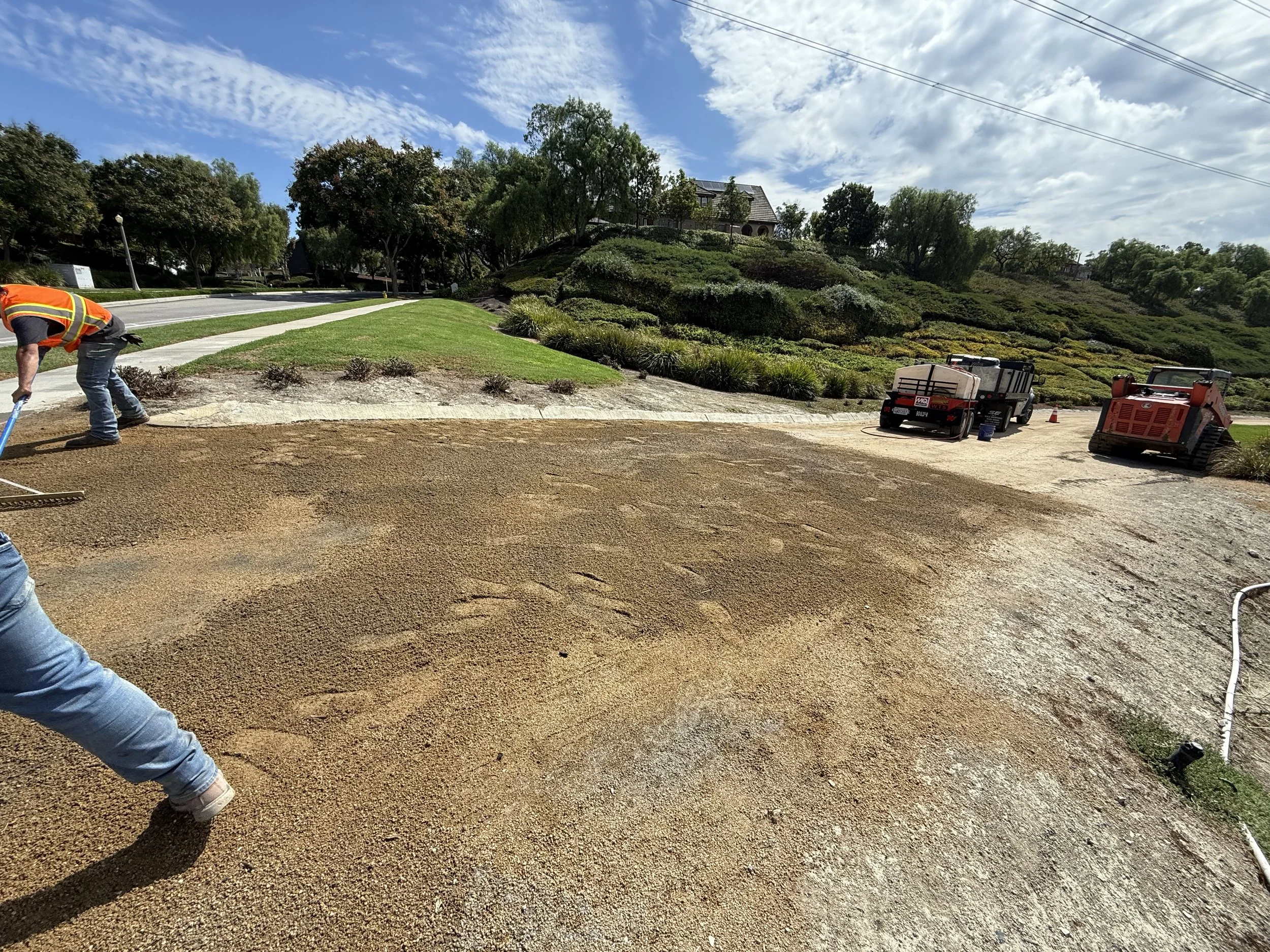 Construction workers installing a gravel pathway on a hillside with lush greenery and structures in the background, under a blue sky with scattered clouds.