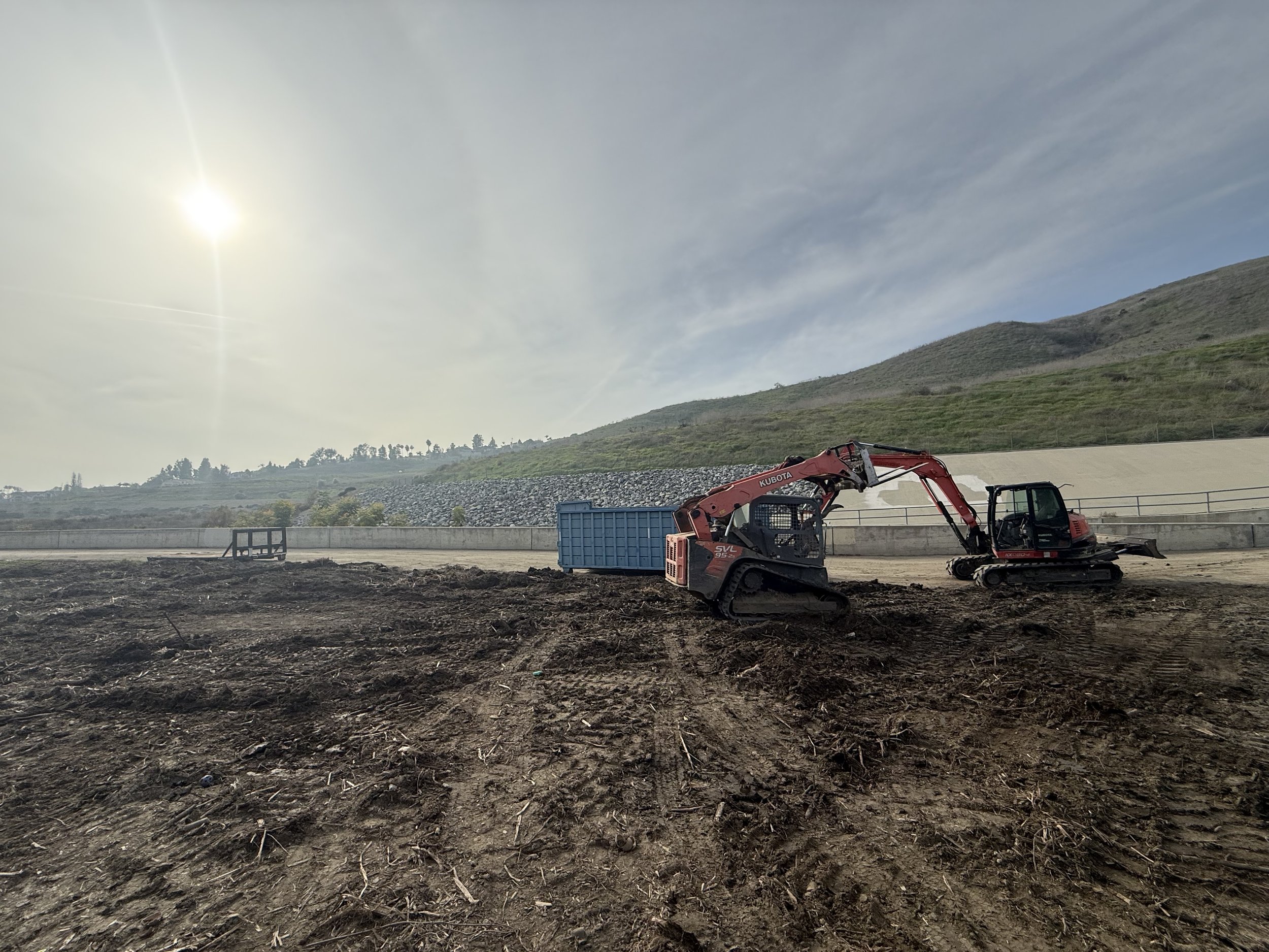 Construction site with tracked excavator digging soil and a blue container in the background. Hills and cloudy sky with the sun shining overhead.