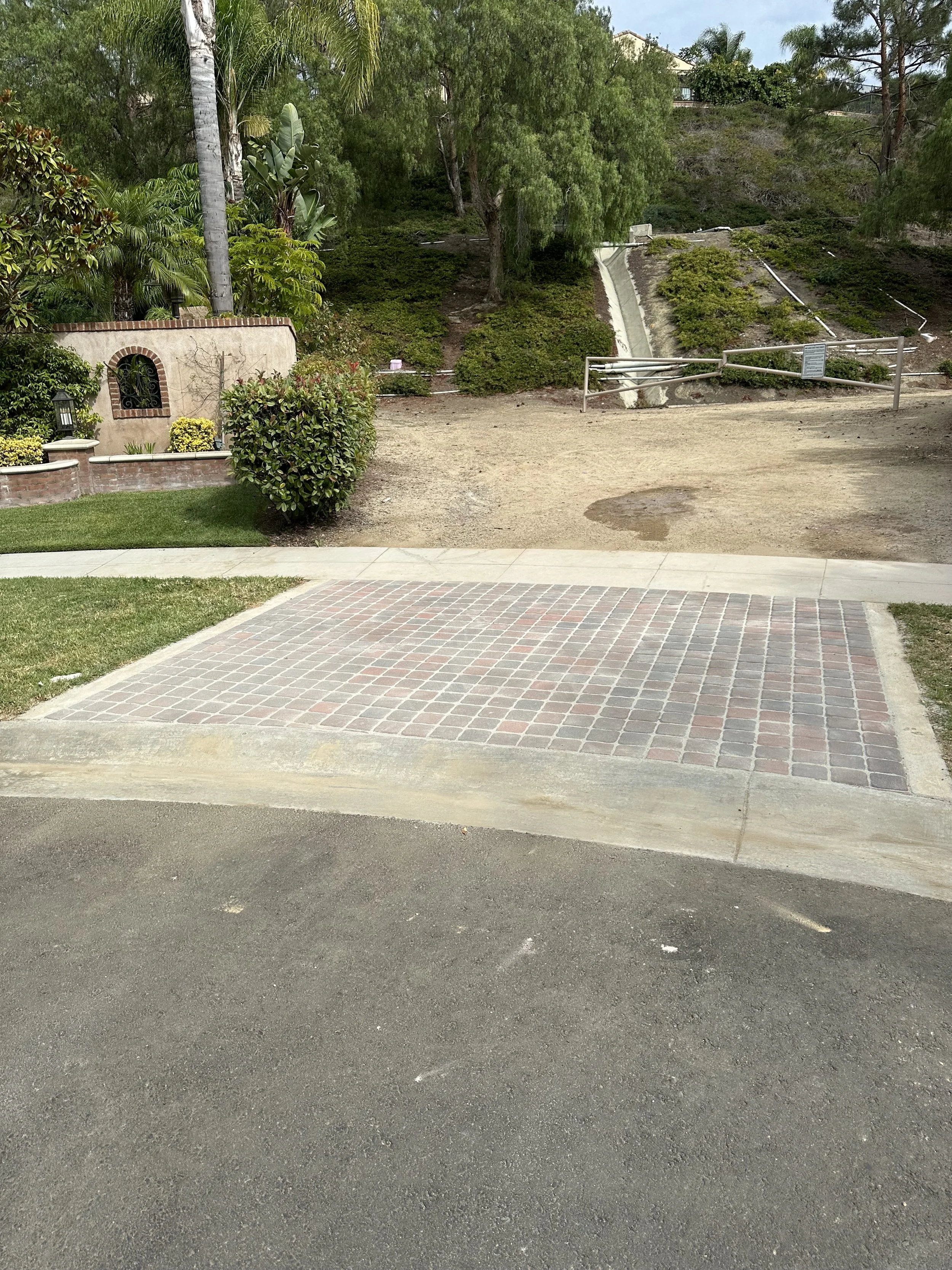 A paved parking spot with red and gray bricks, adjacent to a grassy area and a dirt path leading up a hill with greenery, trees, and a small stone wall on the left side.