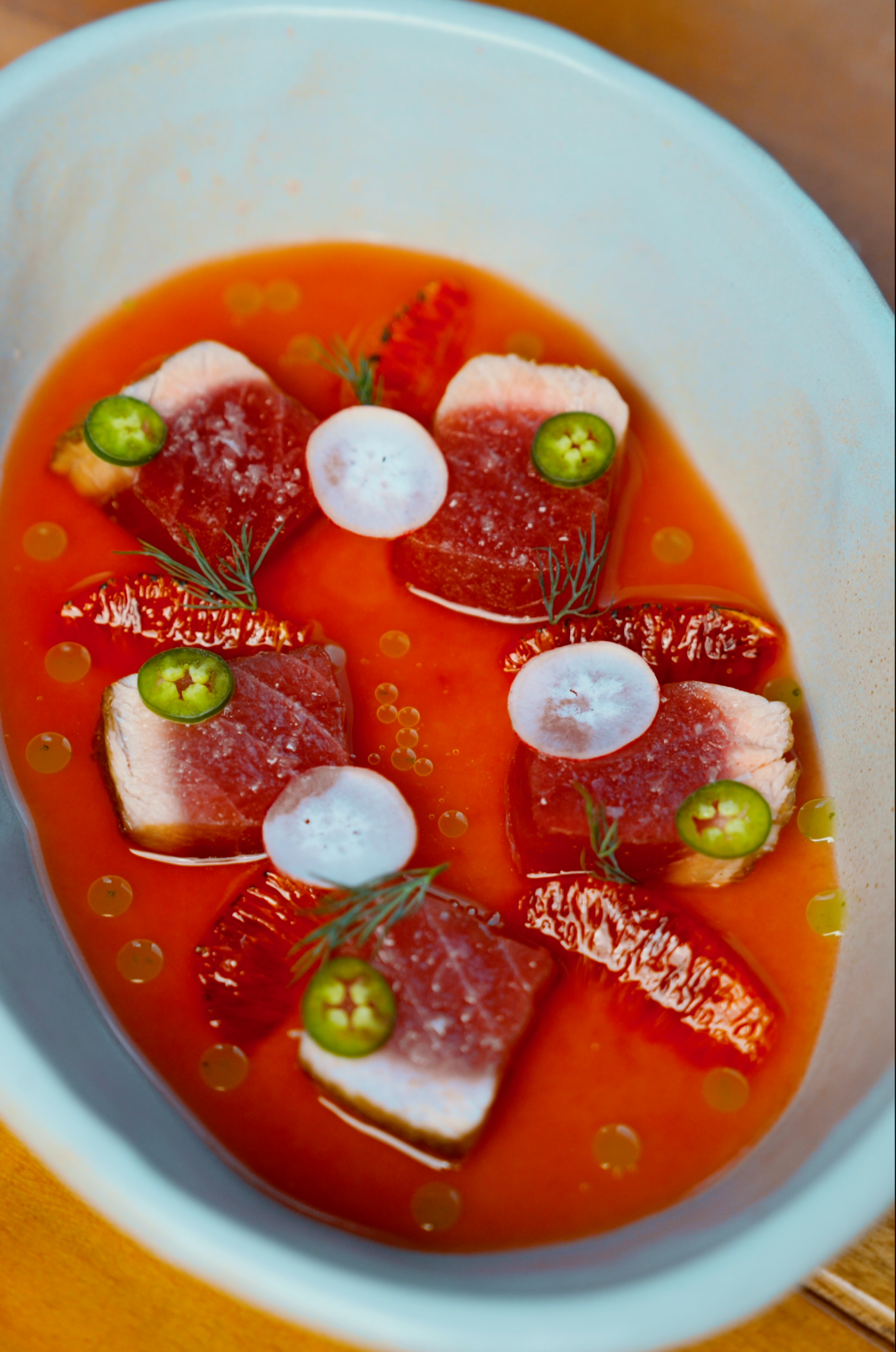 A bowl of cold tomato soup garnished with slices of white cheese, green chili slices, fresh herbs, and pieces of tomato.