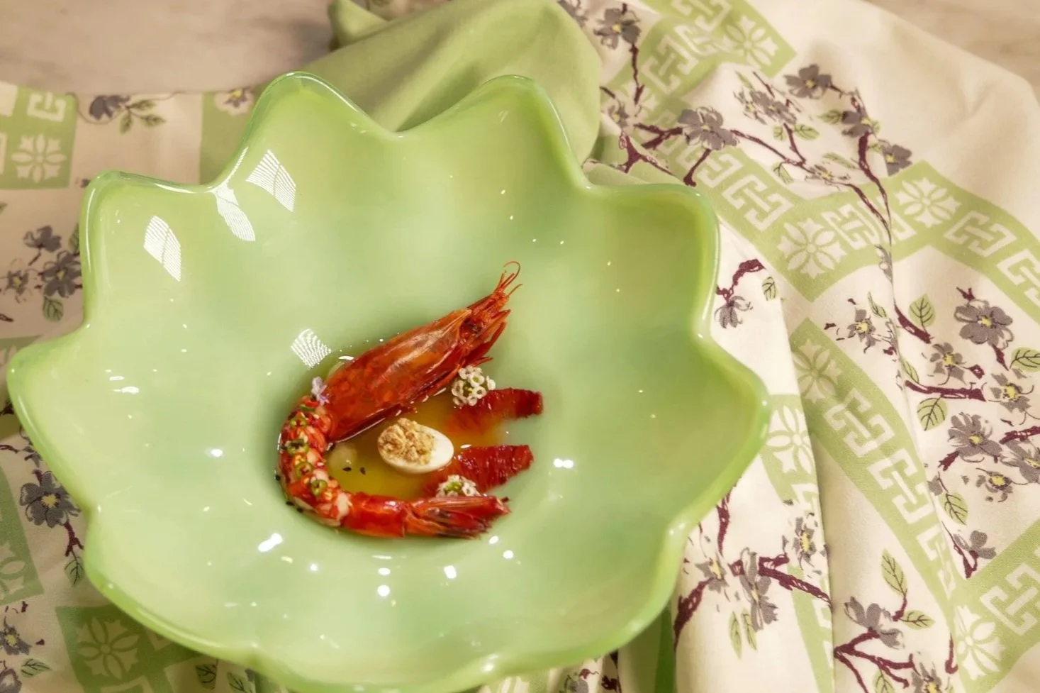 A green flower-shaped bowl containing a cooked shrimp, a half-boiled egg, and some sauce or red seasoning, set on a patterned cloth.